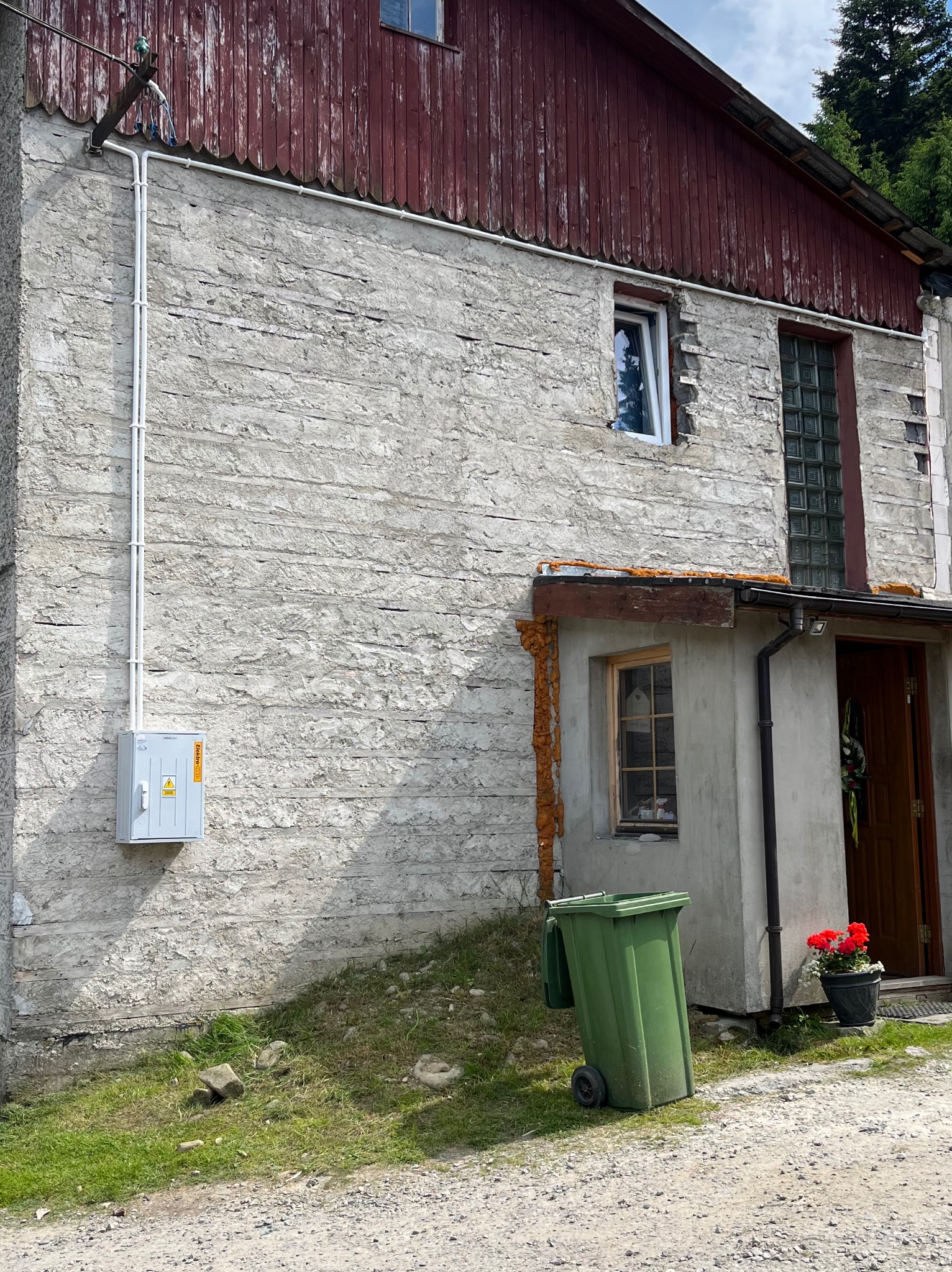 Szara skrzynka elektryczna z białymi rurami na szarej, chropowatej ścianie budynku z czerwonymi elementami drewnianymi. Widoczny fragment trawnika i ścieżki.