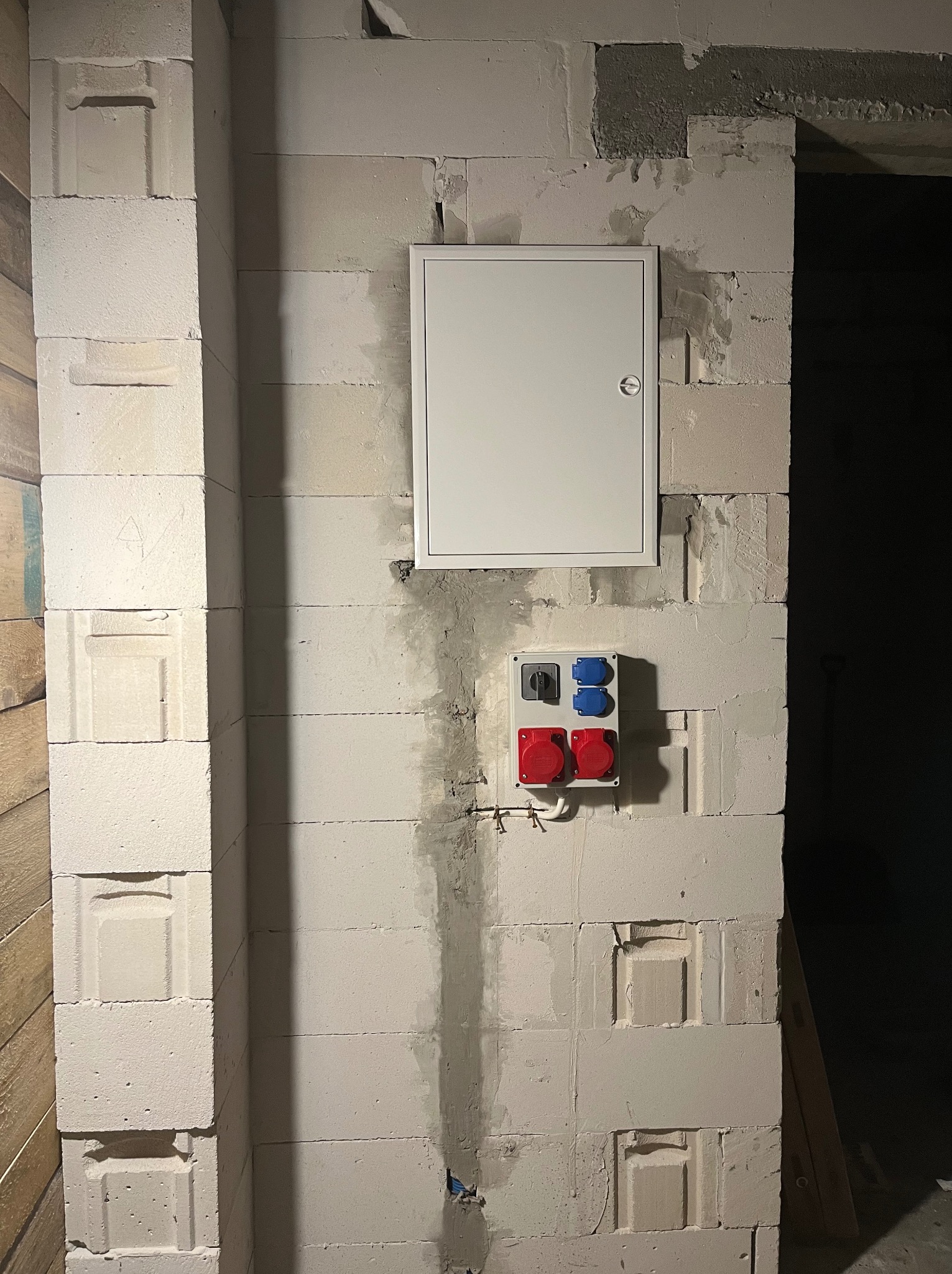 Biała skrzynka elektryczna i gniazda siłowe (czerwone i niebieskie) zamontowane na surowej ścianie z betonu komórkowego wewnątrz budynku.