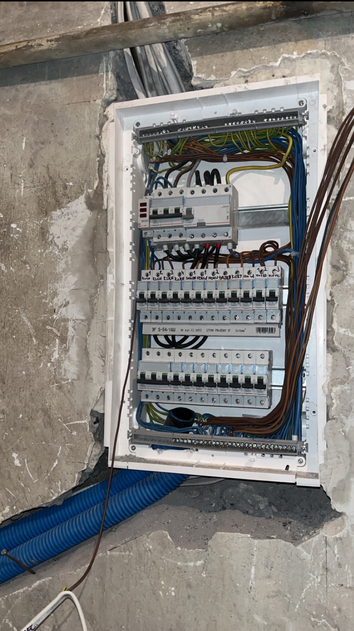 Rozdzielnia elektryczna z licznymi bezpiecznikami, zamontowana w surowej ścianie. Widoczne kable i niebieskie rury instalacyjne. Etap prac wykończeniowych.