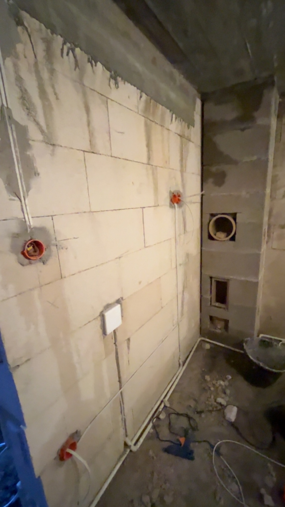 Ściana z betonu komórkowego w trakcie prac instalacyjnych: widoczne puszki elektryczne, przewody i otwór wentylacyjny. Surowy stan wykończenia wnętrza.