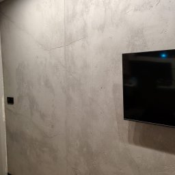 DEKOFAKTURA ADAM ŁABĘCKI - Ściana z szarą, dekoracyjną fakturą przypominającą beton, z delikatnymi spękaniami. Na ścianie wisi czarny telewizor. Widoczny fragment podłogi i listwy przypodłogowej.