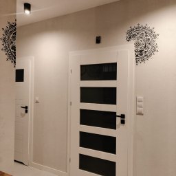 DEKOFAKTURA ADAM ŁABĘCKI - Korytarz z dwiema białymi drzwiami, dekoracyjnymi naklejkami na ścianach i nowoczesnym oświetleniem sufitowym. Widoczna faktura ściany i minimalistyczny design.