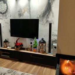 DEKOFAKTURA ADAM ŁABĘCKI - Elegancki salon z dekoracyjną ścianą w odcieniach szarości i złota, podświetloną lampami. Na komodzie świąteczne dekoracje i sprzęt audio. Parkiet i dywan dopełniają wystrój.