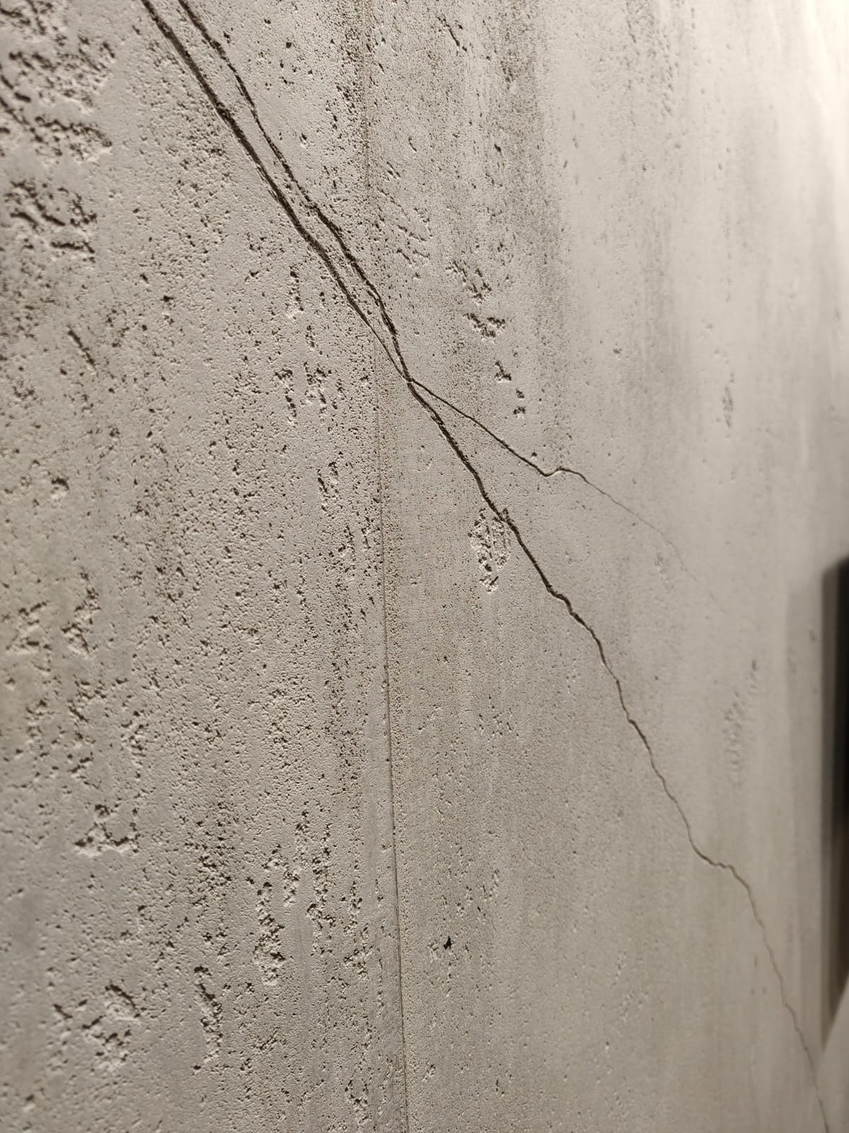 Pęknięcie na szpachlowanej ścianie z widoczną fakturą, biegnące ukośnie od góry do dołu. Szpachlowanie w odcieniach szarości, nierówna powierzchnia.