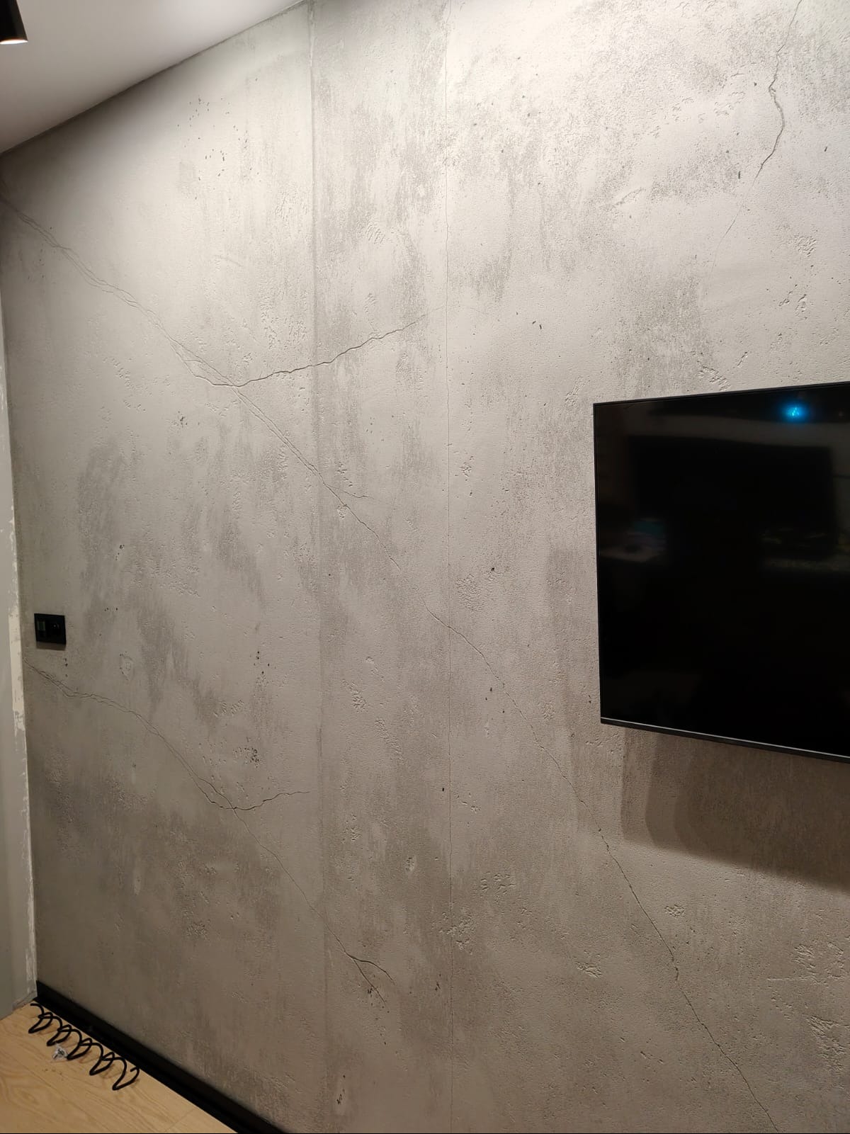Ściana z szarą, dekoracyjną fakturą przypominającą beton, z delikatnymi spękaniami. Na ścianie wisi czarny telewizor. Widoczny fragment podłogi i listwy przypodłogowej.