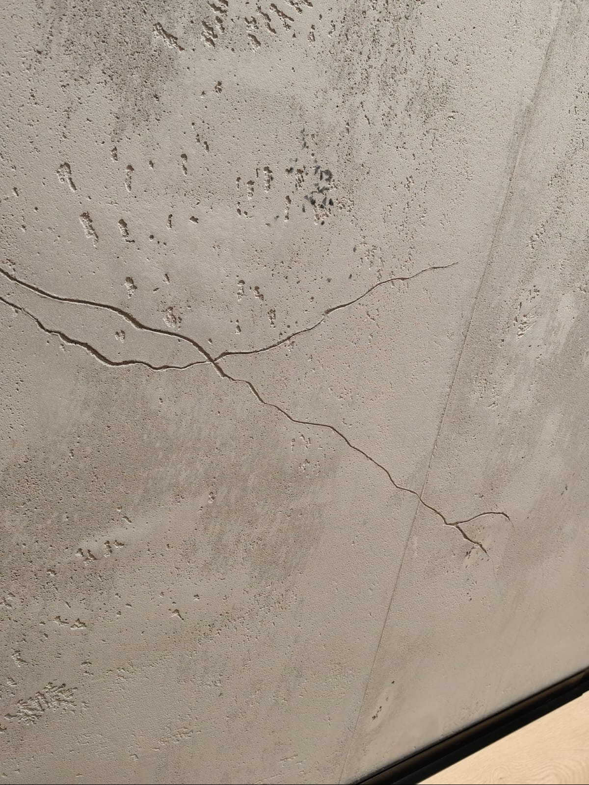 Szara, chropowata powierzchnia ściany z widocznymi pęknięciami i nierównościami, ukazująca strukturę tynku dekoracyjnego w zbliżeniu. Widoczna linia łączenia płyt.