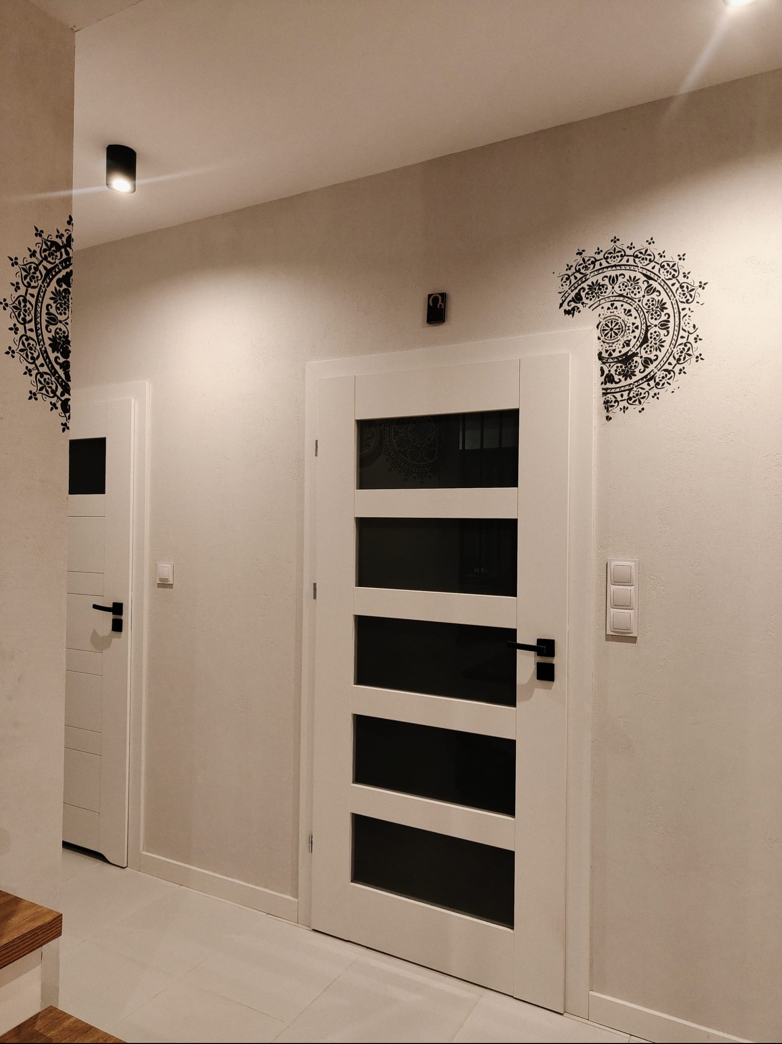 Korytarz z dwiema białymi drzwiami, dekoracyjnymi naklejkami na ścianach i nowoczesnym oświetleniem sufitowym. Widoczna faktura ściany i minimalistyczny design.