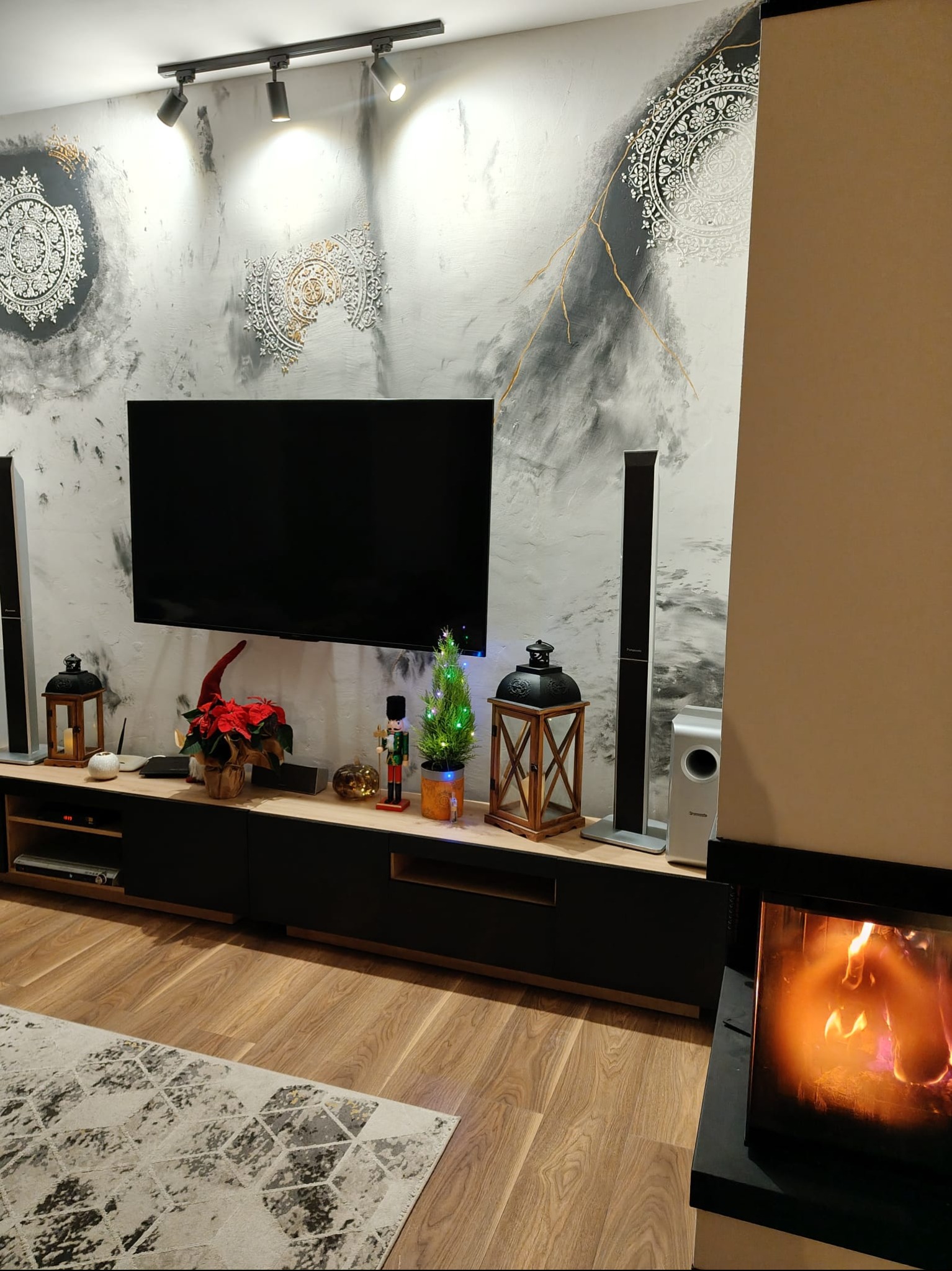 Elegancki salon z dekoracyjną ścianą w odcieniach szarości i złota, podświetloną lampami. Na komodzie świąteczne dekoracje i sprzęt audio. Parkiet i dywan dopełniają wystrój.