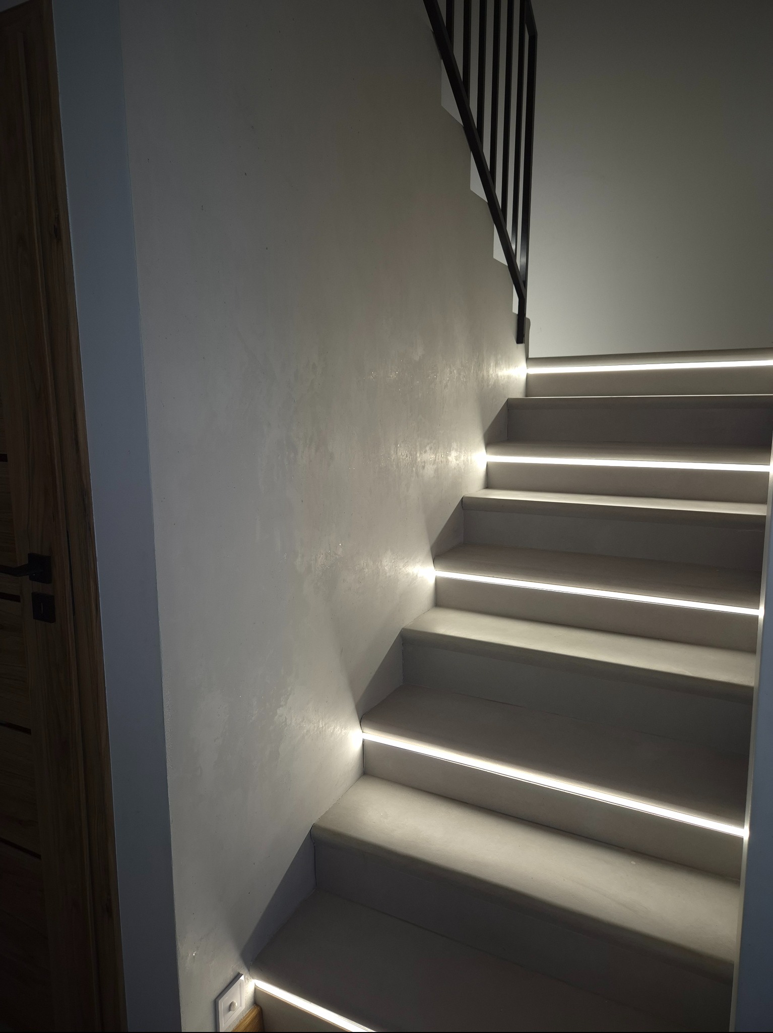 Schody z oświetleniem LED wzdłuż stopni, szara ściana z efektem dekoracyjnym w Owieczkach. Minimalistyczna balustrada i fragment drewnianych drzwi.