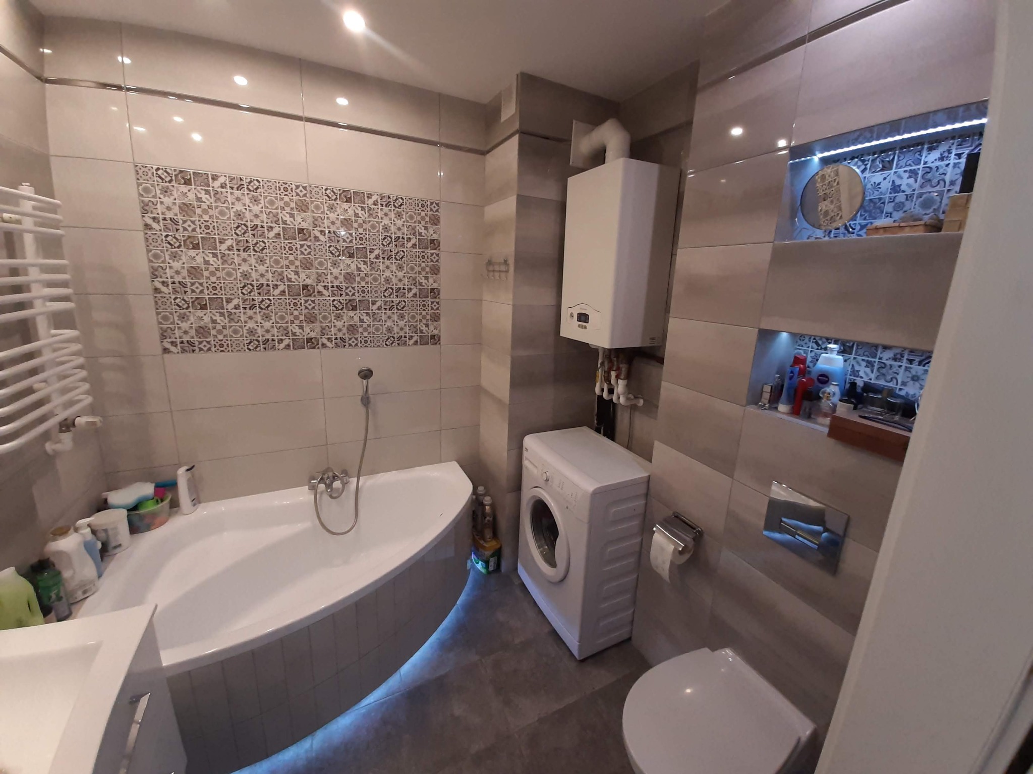 Nowoczesna łazienka z wanną narożną, pralką i podświetlanymi półkami. Beżowe płytki, dekoracyjne mozaiki i subtelne oświetlenie tworzą elegancki wystrój.