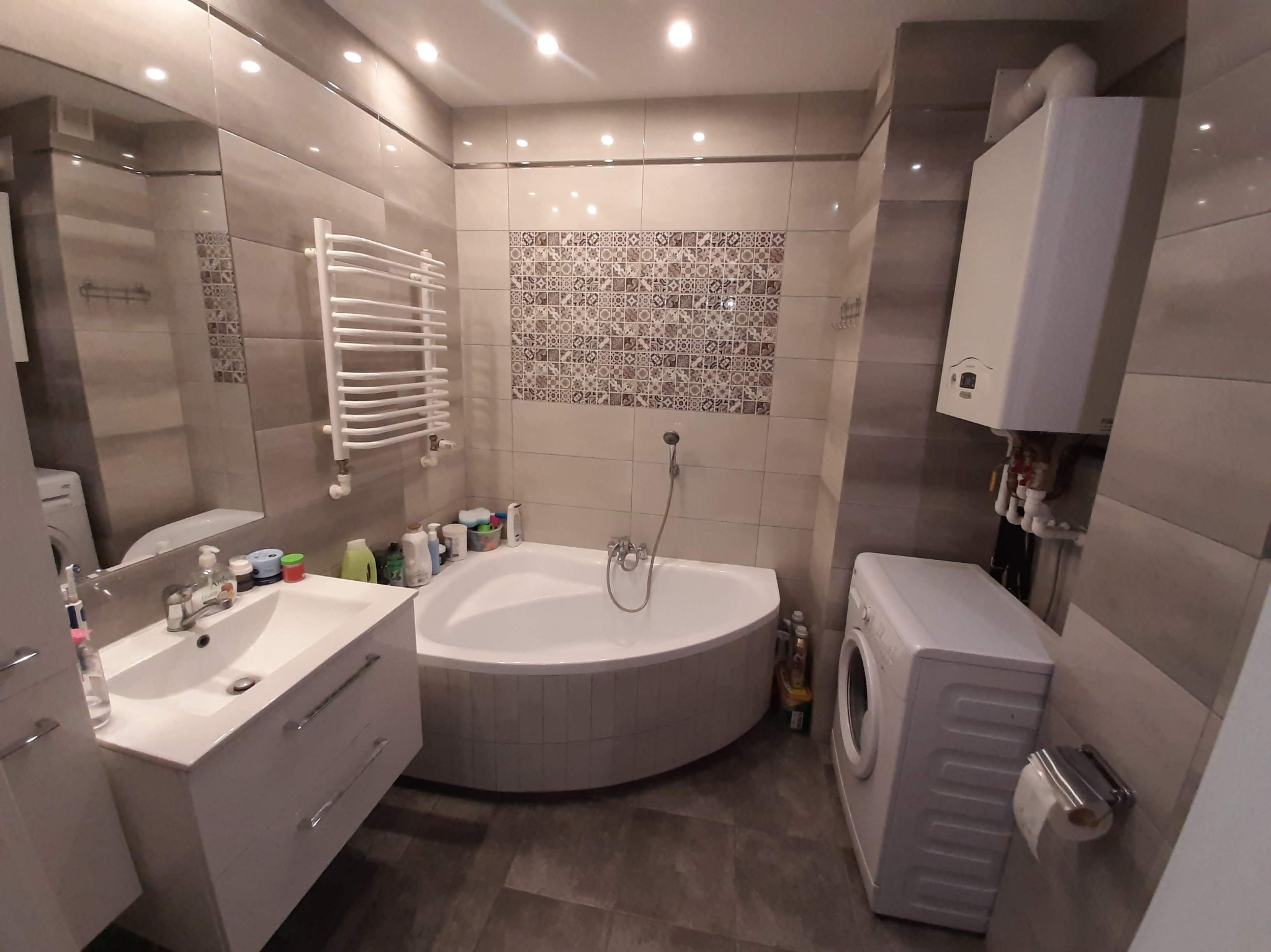 Wykończona łazienka z narożną wanną, umywalką z szafką, grzejnikiem drabinkowym i pralką. Płytki w odcieniach szarości i beżu. Oświetlenie sufitowe punktowe.