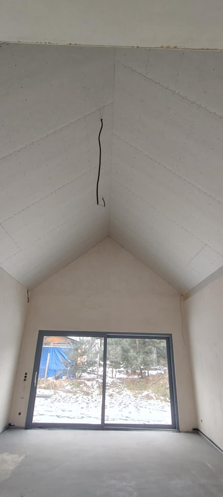 Poddasze z oknem i płytami gipsowymi na skosach dachu, widoczne kable. Ściany wyszpachlowane, podłoga wylewka. Widok na zasypany śniegiem ogród.