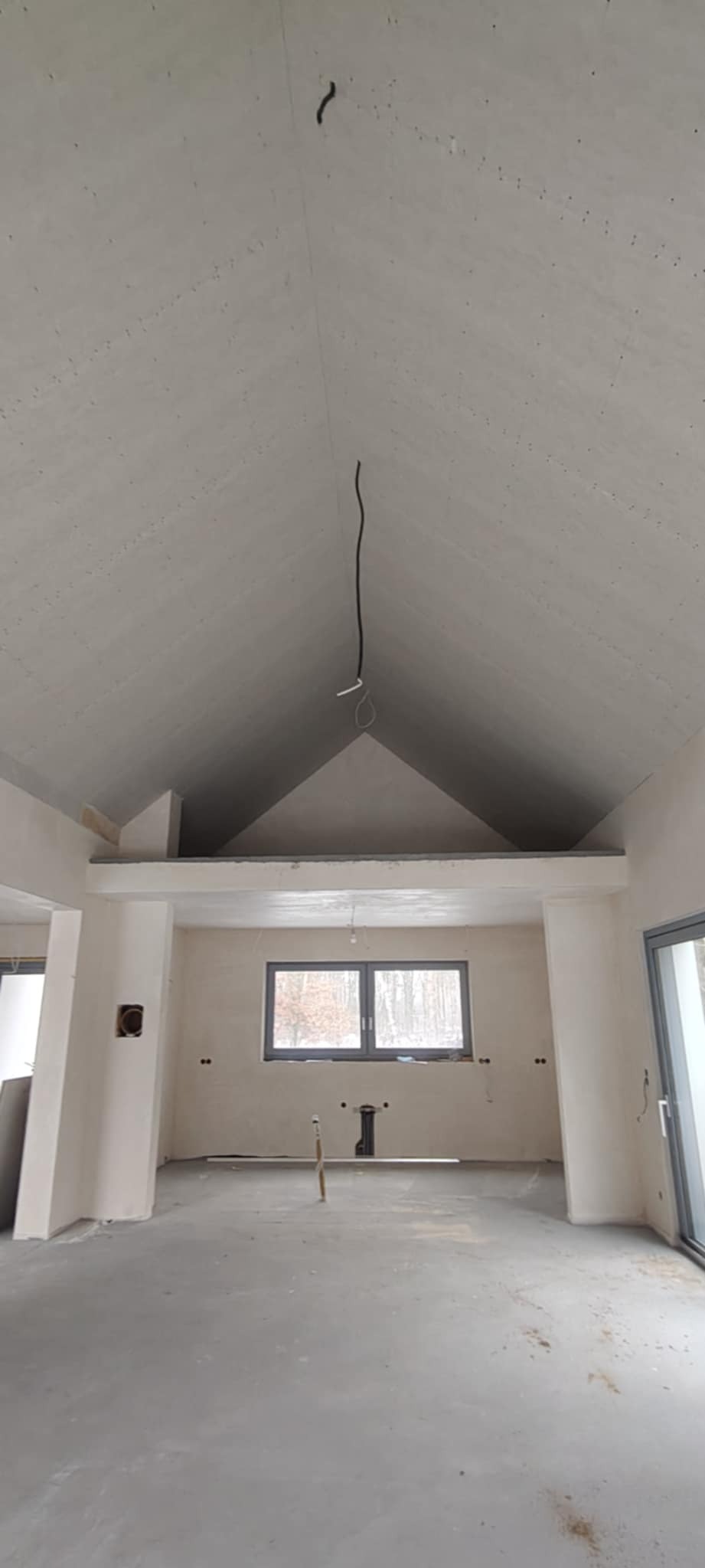 Wnętrze w trakcie wykańczania: sufit z płyt karton-gips, okno z widokiem na las, widoczne instalacje. Minimalistyczny styl, surowe ściany, przygotowanie do dalszych prac.