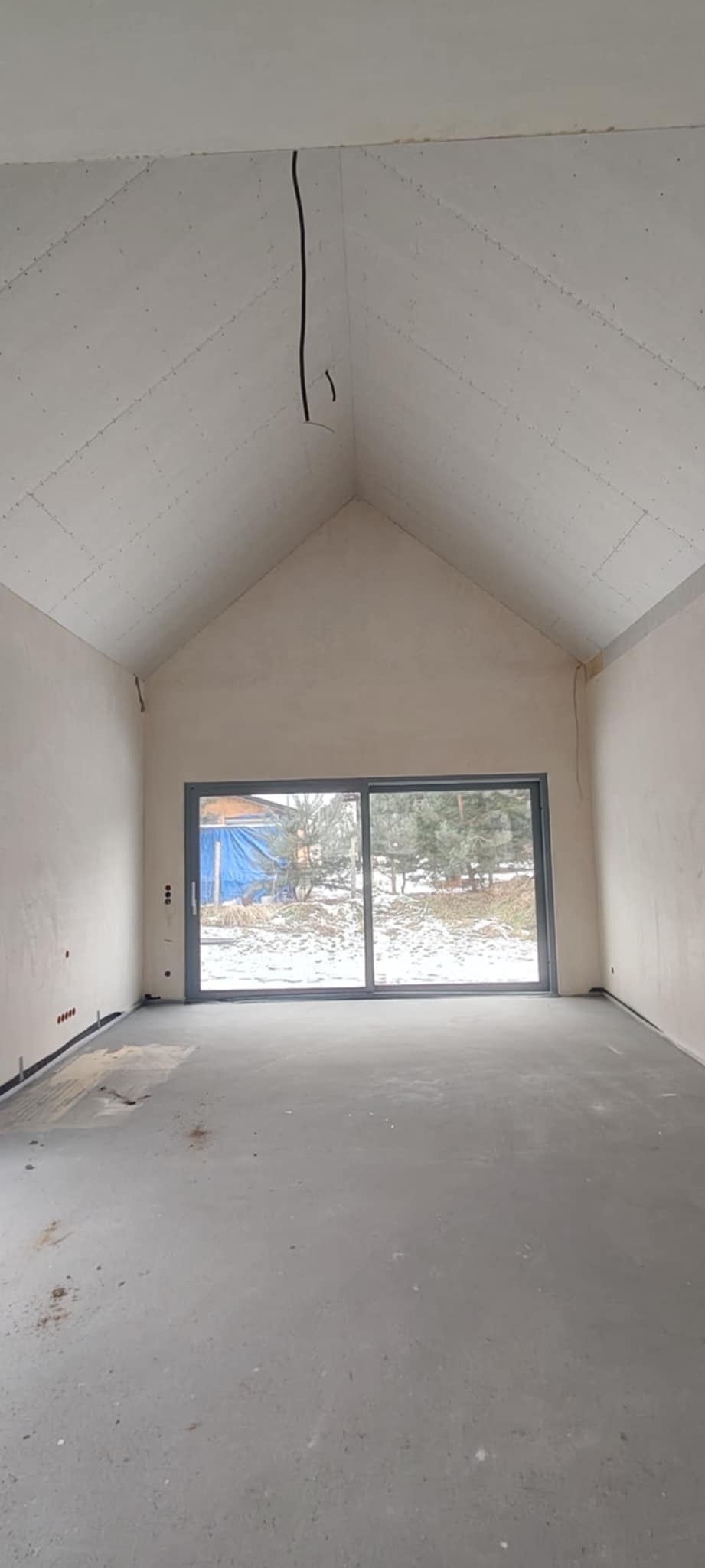 Wnętrze w trakcie wykańczania: sufit z płyt karton-gips, ściany po szpachlowaniu, duże okno przesuwne z widokiem na zimowy krajobraz. Widoczne kable elektryczne.