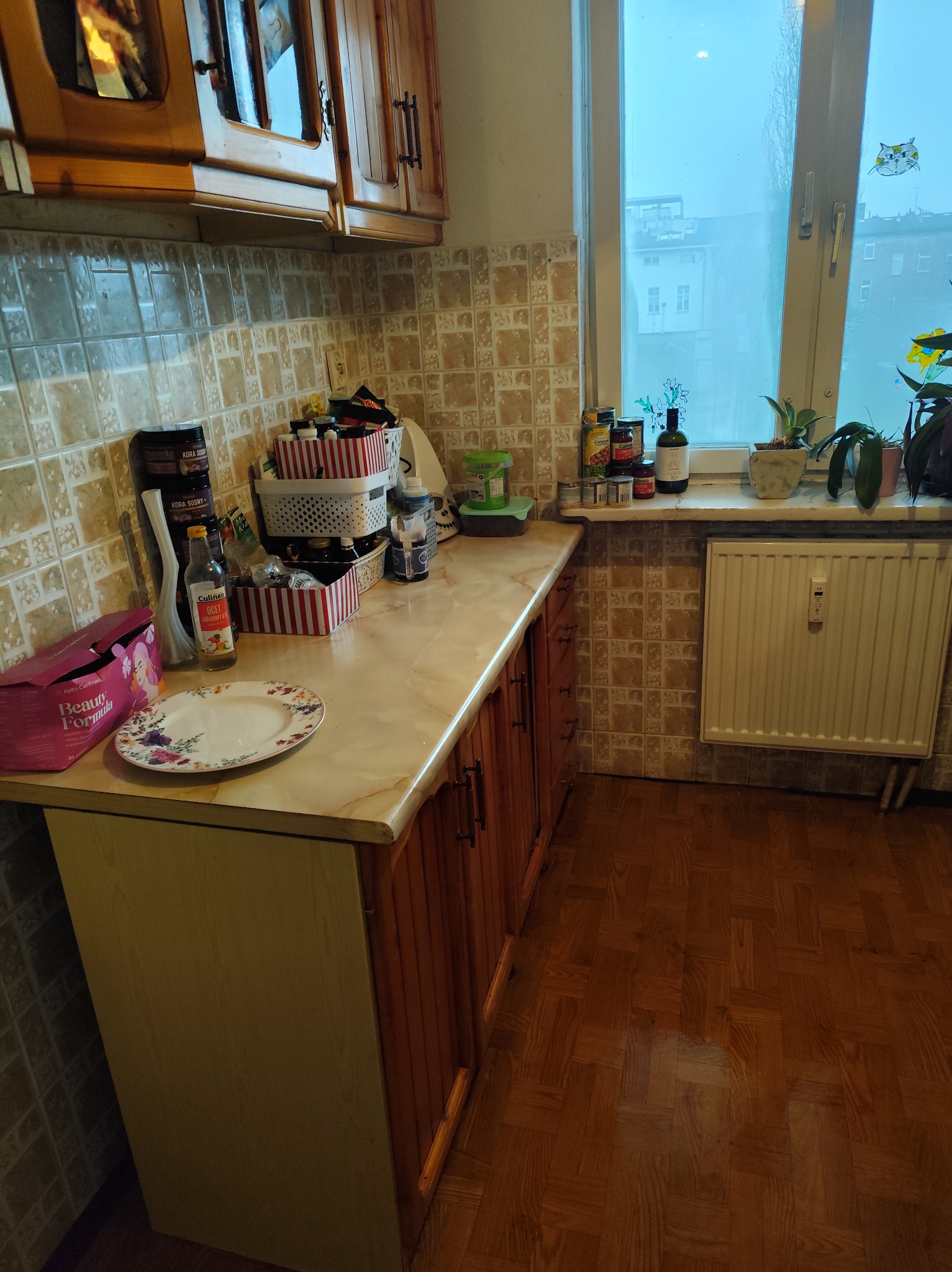 Ujęcie blatu kuchennego z różnymi przedmiotami, w tle okno z naklejką kota. Widoczne płytki na ścianie i drewniana podłoga. Ustawienie przed sprzątaniem.