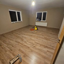 Sebastian Wojtanowski - Pokój po malowaniu ścian na beżowo, z drewnianą podłogą, widoczne narzędzia malarskie i odkurzacz. Dwa okna z białymi ramami w tle.