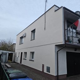 Sebastian Wojtanowski - Dwupiętrowy dom po renowacji elewacji w odcieniach beżu, z balkonem i anteną na dachu, widziany z poziomu chodnika z kostki brukowej. Widoczne kosze na śmieci.