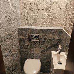 Sebastian Wojtanowski - Wykończona toaleta z szarą, marmurkową okładziną ścian i podłogi. Widoczna biała miska WC, umywalka z szafką i fragment ściany z surowym wykończeniem.