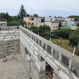 Sebastian Wojtanowski - Wznoszenie ścian z bloczków betonu komórkowego na budowie domu jednorodzinnego. Widoczne narzędzia i elementy konstrukcyjne. Tło stanowią zabudowania mieszkalne i zieleń.