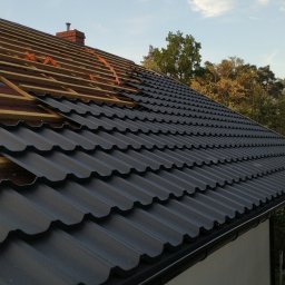 Sebastian Wojtanowski - Częściowo pokryty dach z czarnej blachy trapezowej, widoczne drewniane łaty i komin na tle błękitnego nieba z chmurami i zieleni drzew.