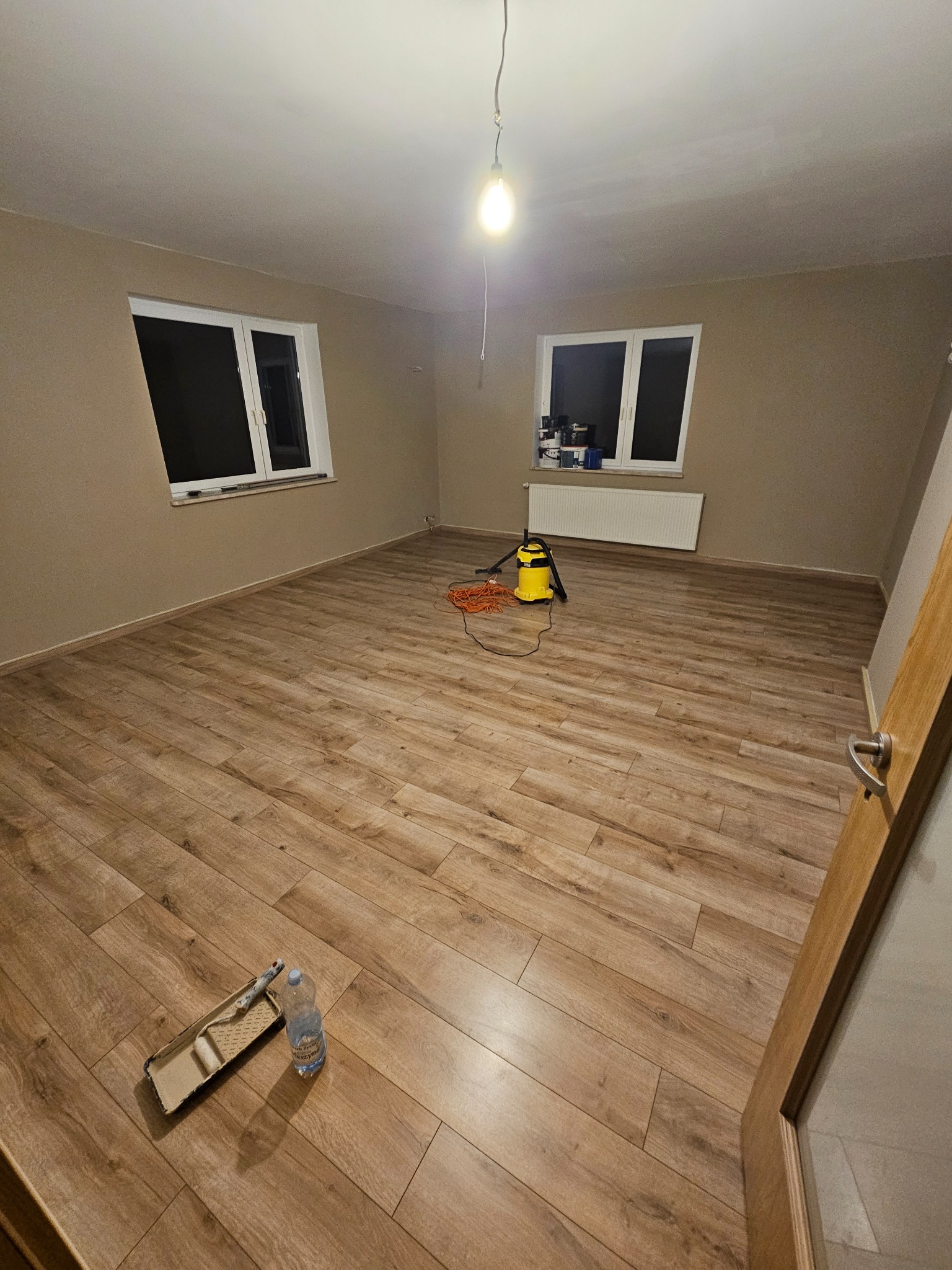 Pokój po malowaniu ścian na beżowo, z drewnianą podłogą, widoczne narzędzia malarskie i odkurzacz. Dwa okna z białymi ramami w tle.