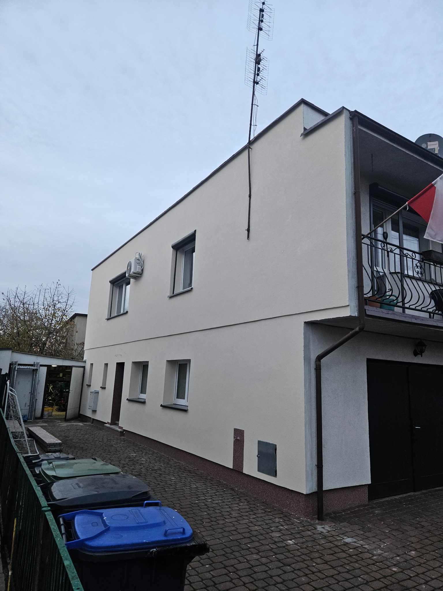 Dwupiętrowy dom po renowacji elewacji w odcieniach beżu, z balkonem i anteną na dachu, widziany z poziomu chodnika z kostki brukowej. Widoczne kosze na śmieci.