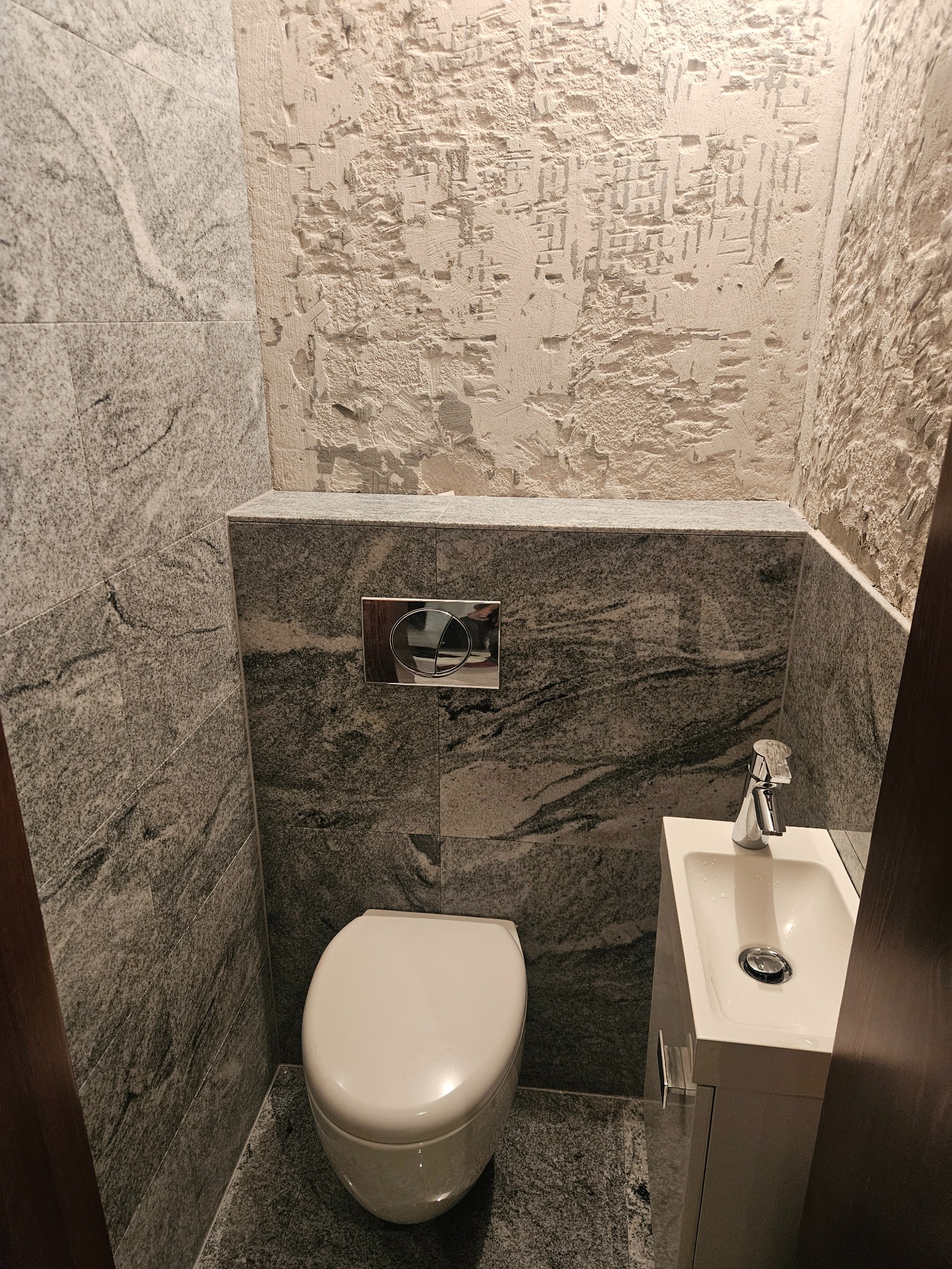 Wykończona toaleta z szarą, marmurkową okładziną ścian i podłogi. Widoczna biała miska WC, umywalka z szafką i fragment ściany z surowym wykończeniem.