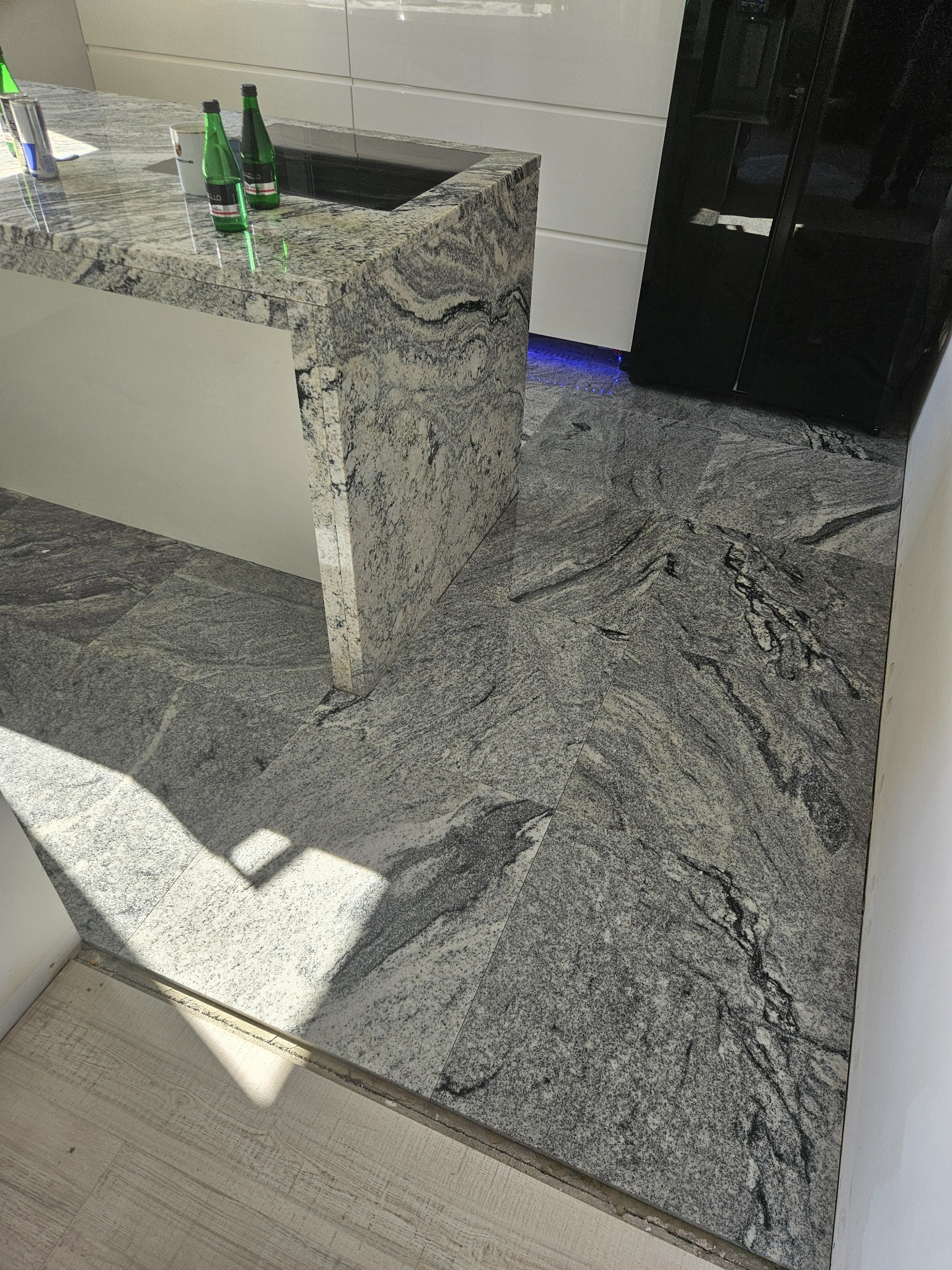Kuchnia z blatem i podłogą z szarego, wzorzystego kamienia. Widoczne butelki na blacie i fragment czarnej lodówki. Nowoczesny design z akcentem na naturalny kamień.