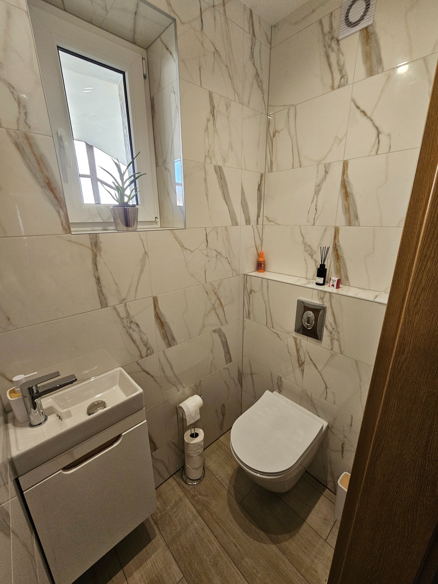 Nowoczesna toaleta z marmurowymi płytkami, wiszącą miską WC i umywalką z szafką. Na półce dekoracje i dyfuzor zapachowy. Podłoga imitująca drewno.