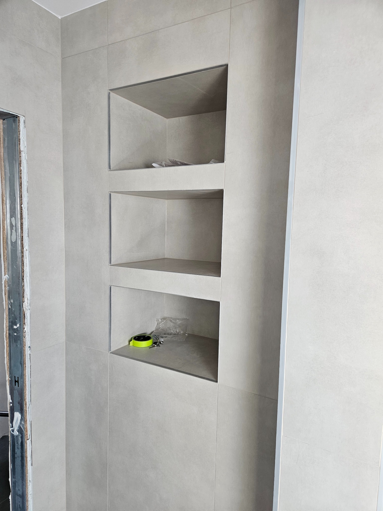 Wykończona wnęka z półkami w łazience, wyłożona jasnymi płytkami. Na dolnej półce leżą narzędzia. Widoczny fragment ościeżnicy drzwi.