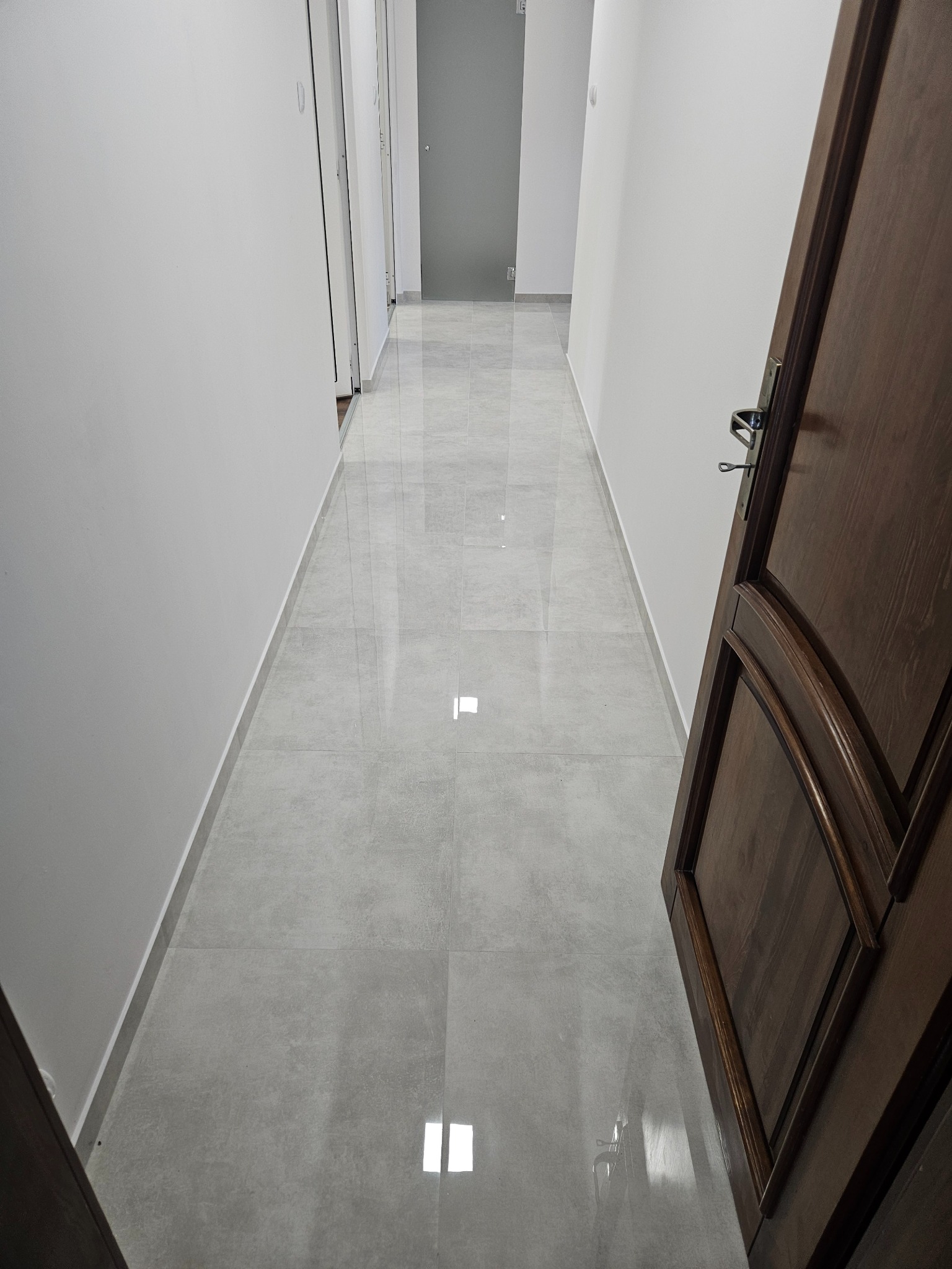 Wykończony korytarz z szarymi, błyszczącymi płytkami podłogowymi i otwartymi, ciemnymi drzwiami po prawej stronie. Ściany pomalowane na biało.
