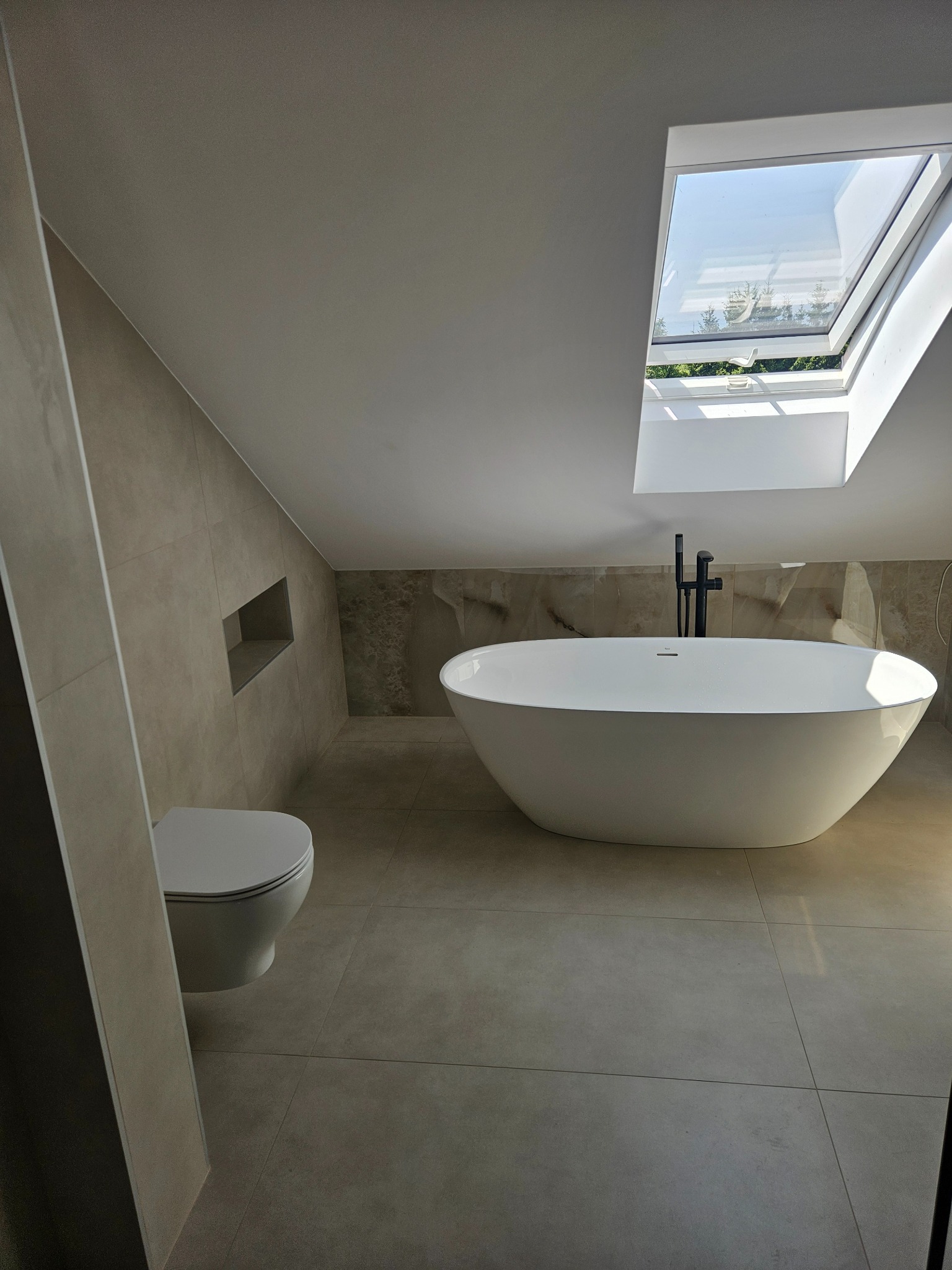 Nowoczesna łazienka na poddaszu z oknem dachowym. Beżowe płytki, wolnostojąca wanna i toaleta w minimalistycznym stylu. Czarna armatura i wnęka w ścianie.