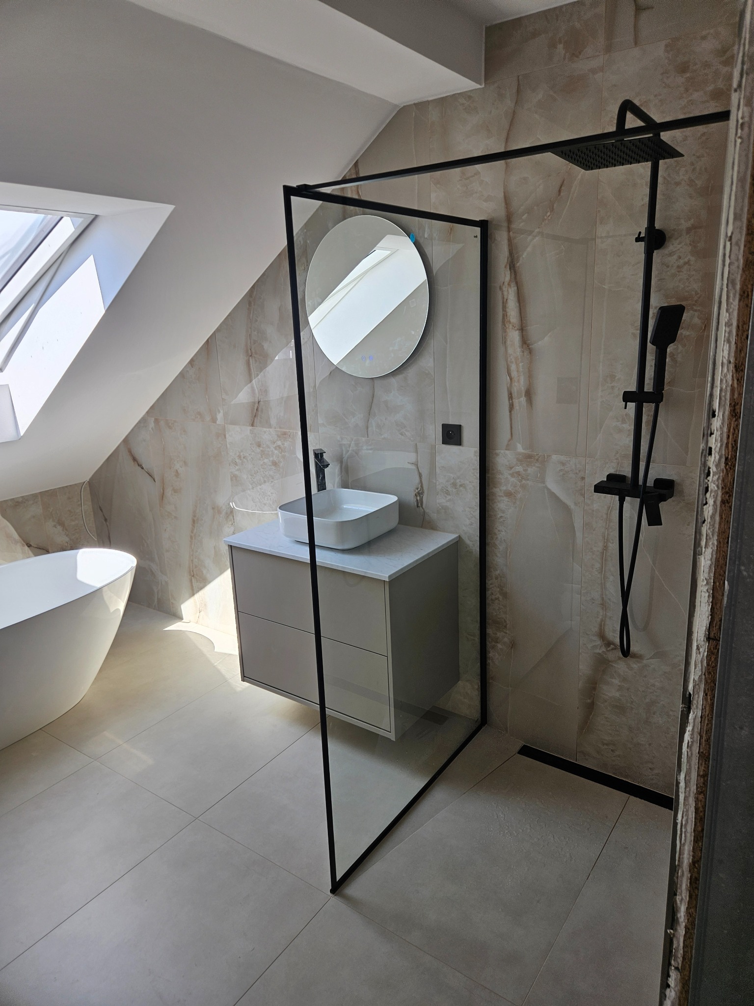 Jasna łazienka na poddaszu ze skośnym sufitem, z wanną, umywalką na szafce i kabiną prysznicową z czarnymi ramami. Beżowe płytki imitujące marmur na ścianach.