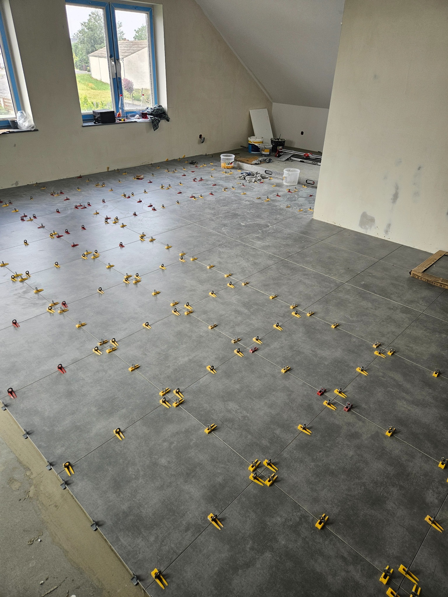 Szare płytki podłogowe w trakcie układania, z systemem poziomowania. Widoczne okna i narzędzia glazurnicze w jasnym, przestronnym pomieszczeniu na poddaszu.