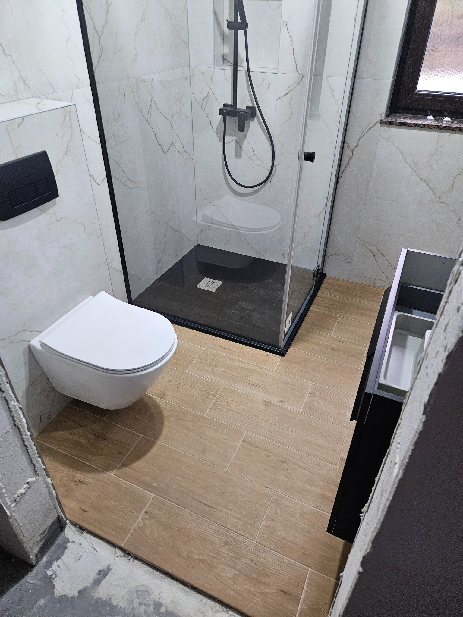 Nowoczesna łazienka po remoncie: kabina prysznicowa typu walk-in z czarnymi profilami, podwieszana toaleta i drewnopodobne płytki podłogowe. Minimalistyczny design.