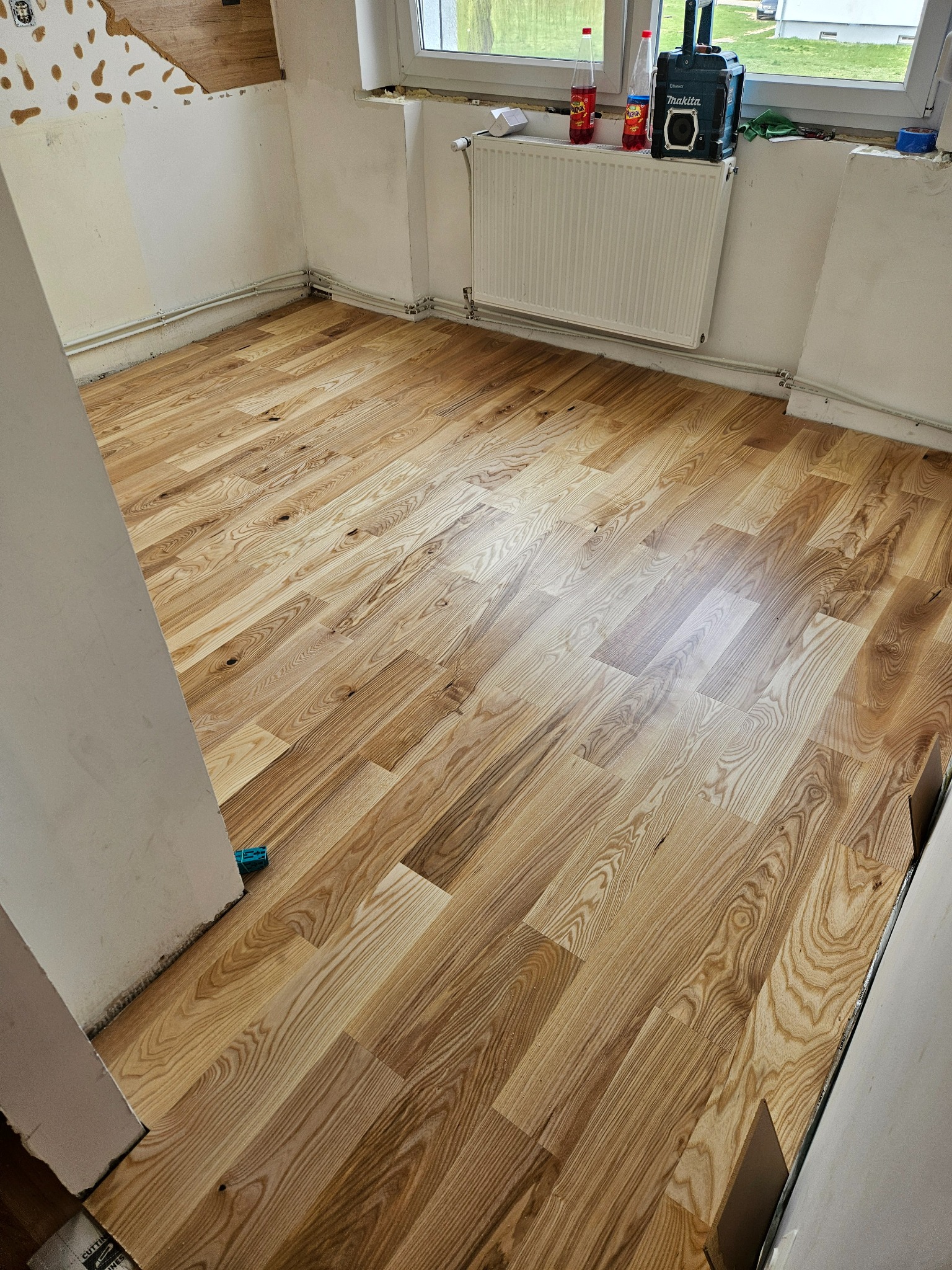 Jasna, drewniana podłoga ułożona w jodełkę w odnawianym pokoju. Widoczne ślady po remoncie na ścianach i narzędzia przy oknie. Podłoga po renowacji.