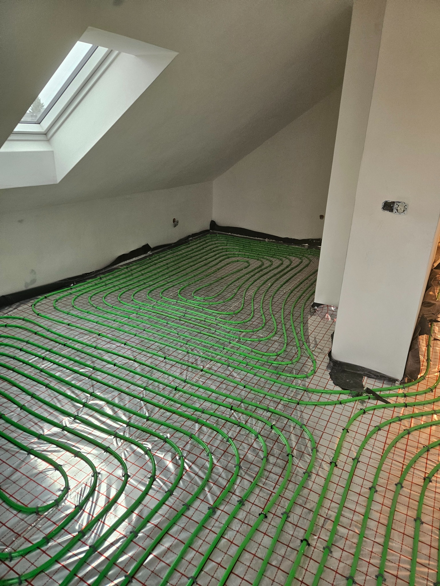 Zielone rury ogrzewania podłogowego ułożone na folii w jasnym poddaszu z oknem dachowym. Widoczne elementy instalacji elektrycznej na ścianie.