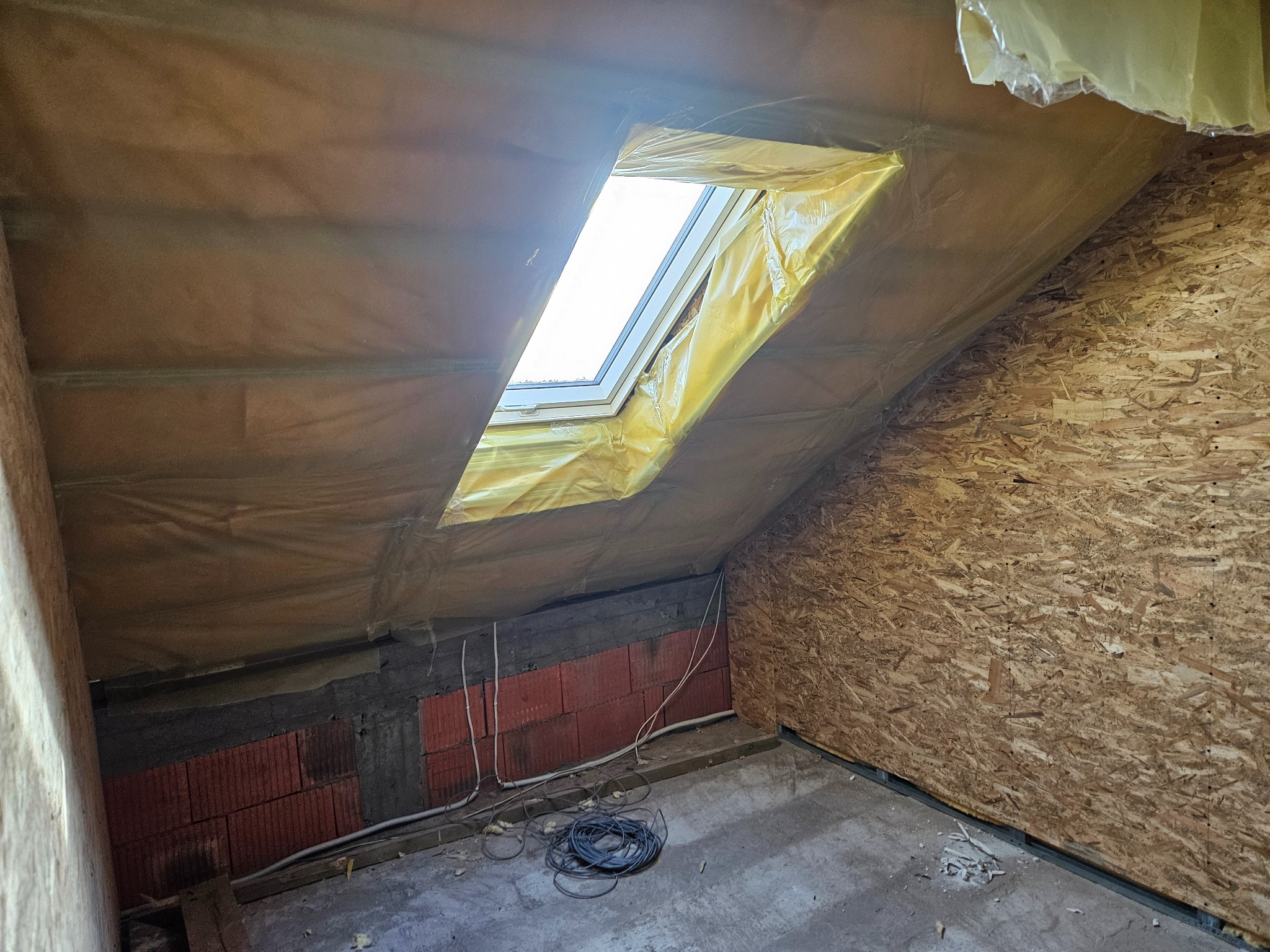 Poddasze w trakcie prac wykończeniowych: izolacja folią okna dachowego, ściana z płyty OSB, fragment ściany z cegły, widoczne przewody elektryczne.