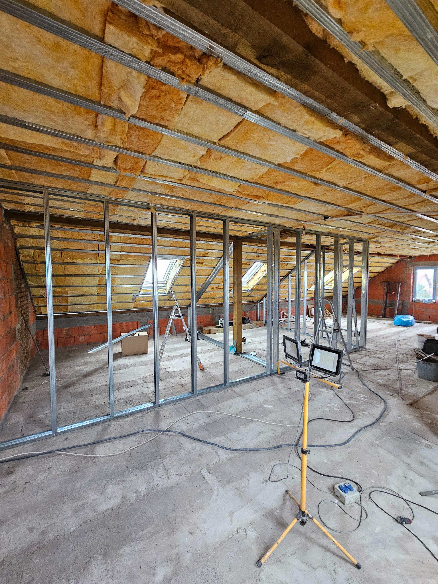 Szkielet ścian działowych z profili stalowych na poddaszu, ocieplenie wełną mineralną, widoczne okno dachowe i lampa budowlana na trójnogu.