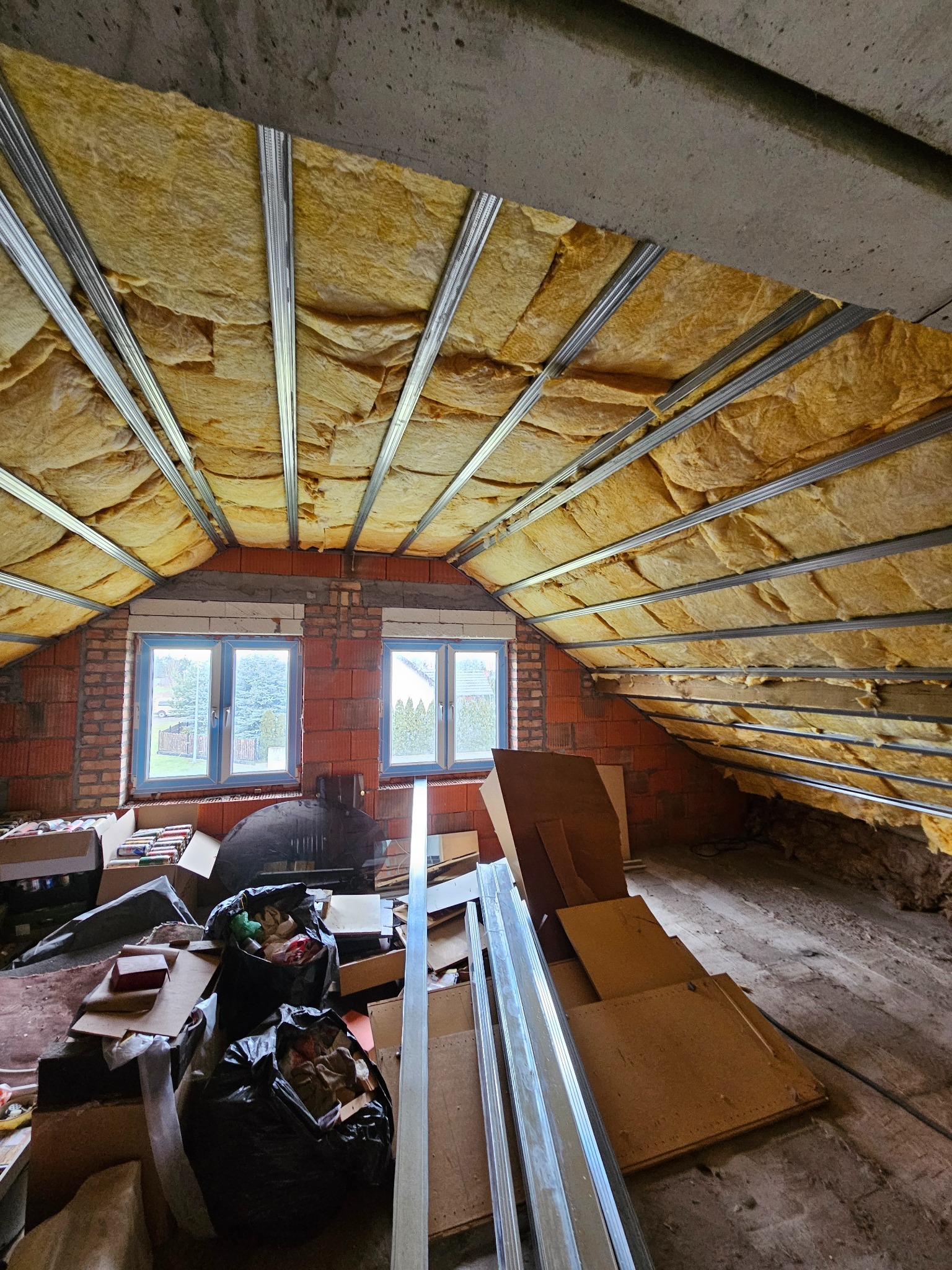 Poddasze w trakcie adaptacji: widoczne ocieplenie wełną mineralną między metalowymi profilami, surowe ściany z cegły i okna. Na podłodze gruz i materiały budowlane.