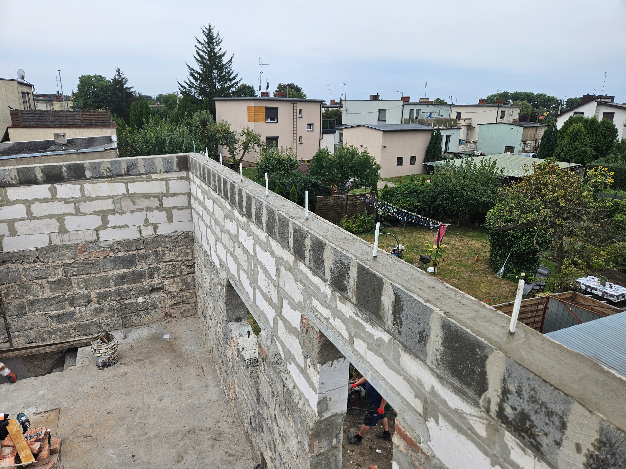 Wznoszenie ścian z bloczków betonu komórkowego na budowie domu jednorodzinnego. Widoczne narzędzia i elementy konstrukcyjne. Tło stanowią zabudowania mieszkalne i zieleń.