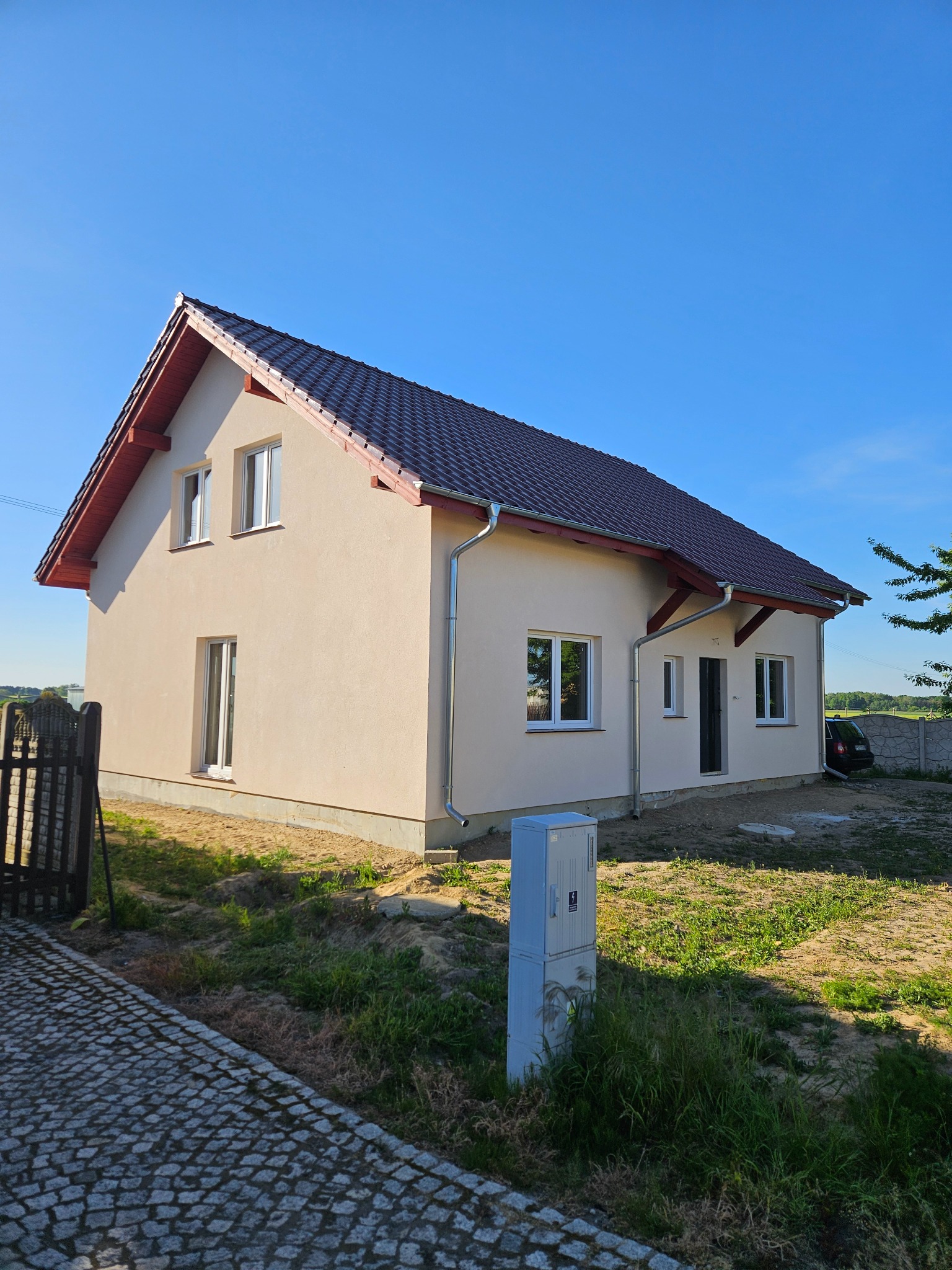 Otynkowany dom jednorodzinny z brązowym dachem i białymi oknami w słoneczny dzień. Widoczne rynny i skrzynka elektryczna. Brukowany chodnik.