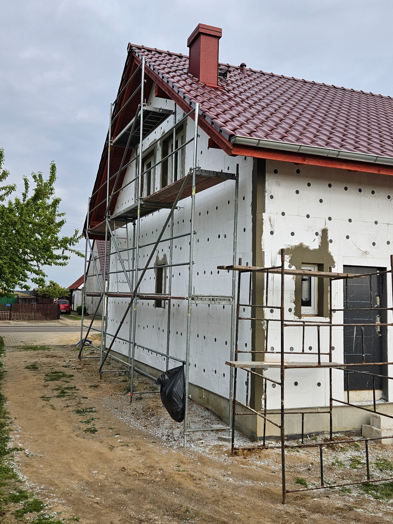 Ocieplanie domu styropianem, widoczne rusztowania i fragment elewacji z nałożonym klejem wokół okna. Czerwona dachówka i komin. Prace w toku.
