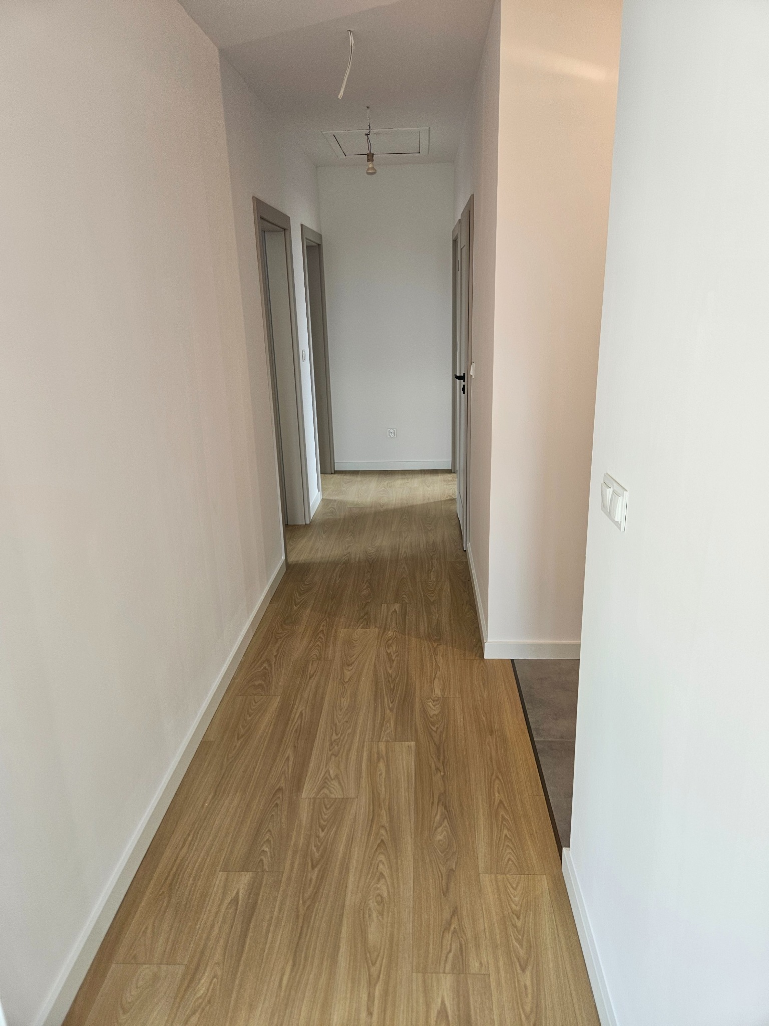 Jasny korytarz po remoncie: drewnopodobna podłoga, świeżo pomalowane białe ściany, szare futryny drzwi. Widoczne detale wykończenia i oświetlenie.