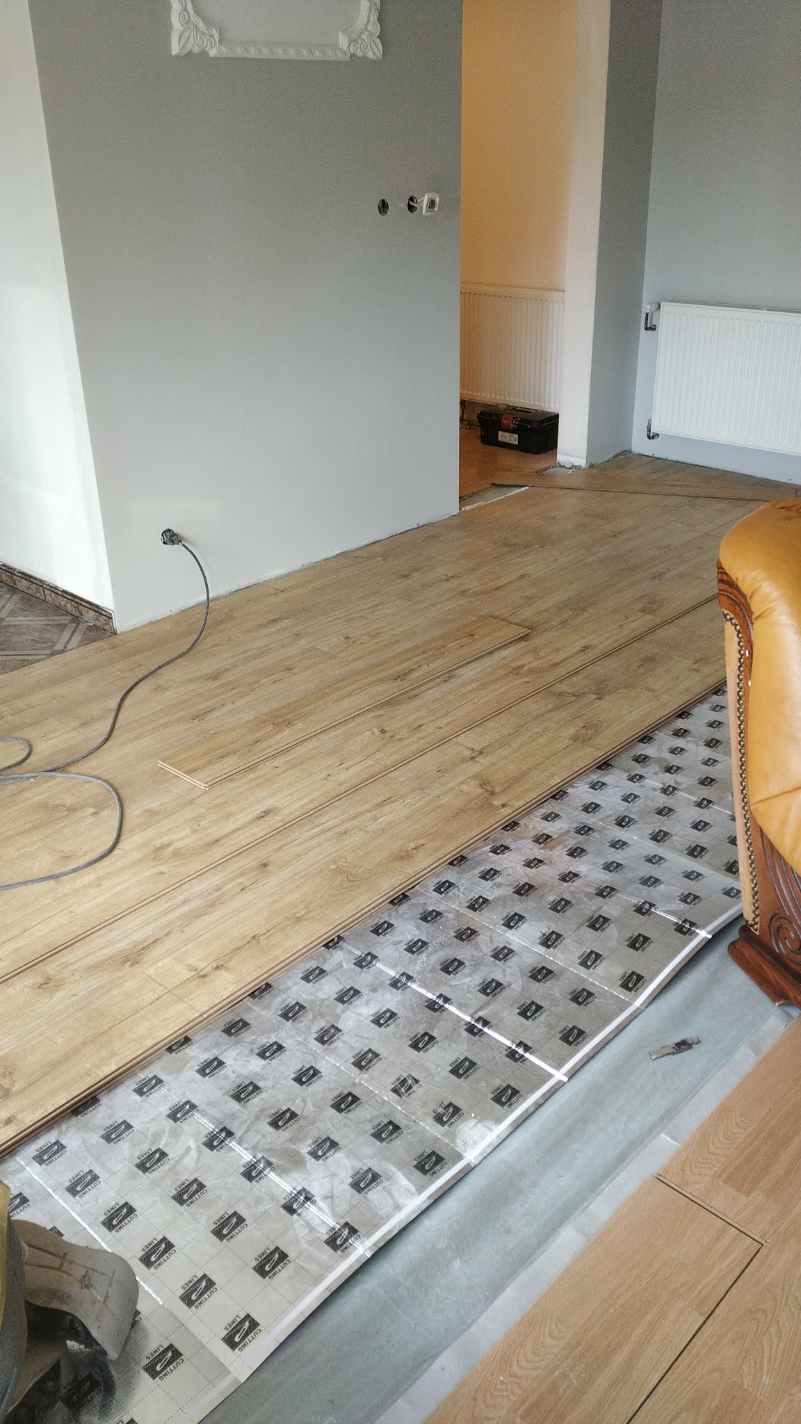 Układanie paneli podłogowych na folii izolacyjnej w jasnym wnętrzu. Częściowo ułożona podłoga, widoczne narzędzia i fragment sofy. Prace wykończeniowe w toku.