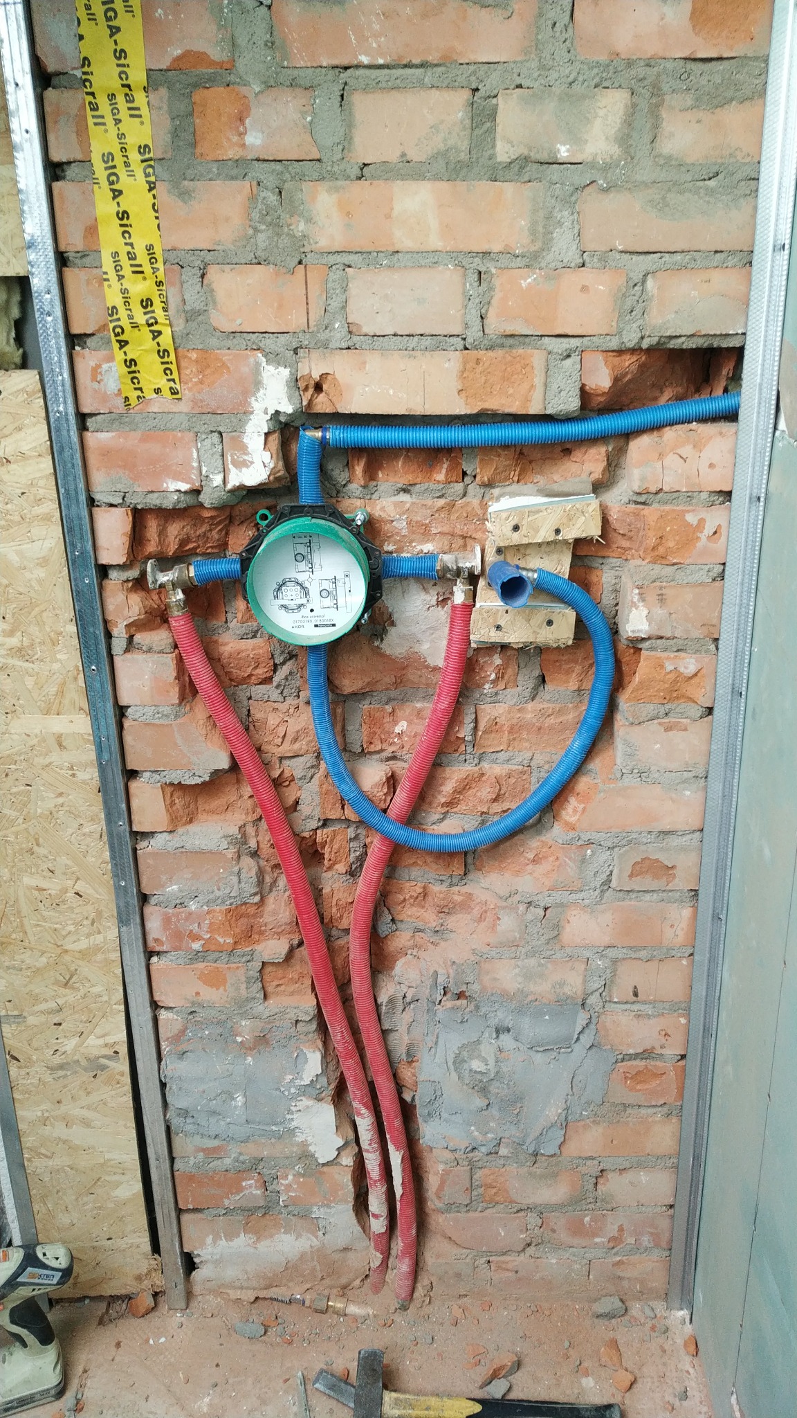 Instalacja wodna w ścianie z czerwonej cegły, z widocznymi rurami w kolorze niebieskim i czerwonym, oraz elementami montażowymi. Widoczne narzędzia na podłodze.
