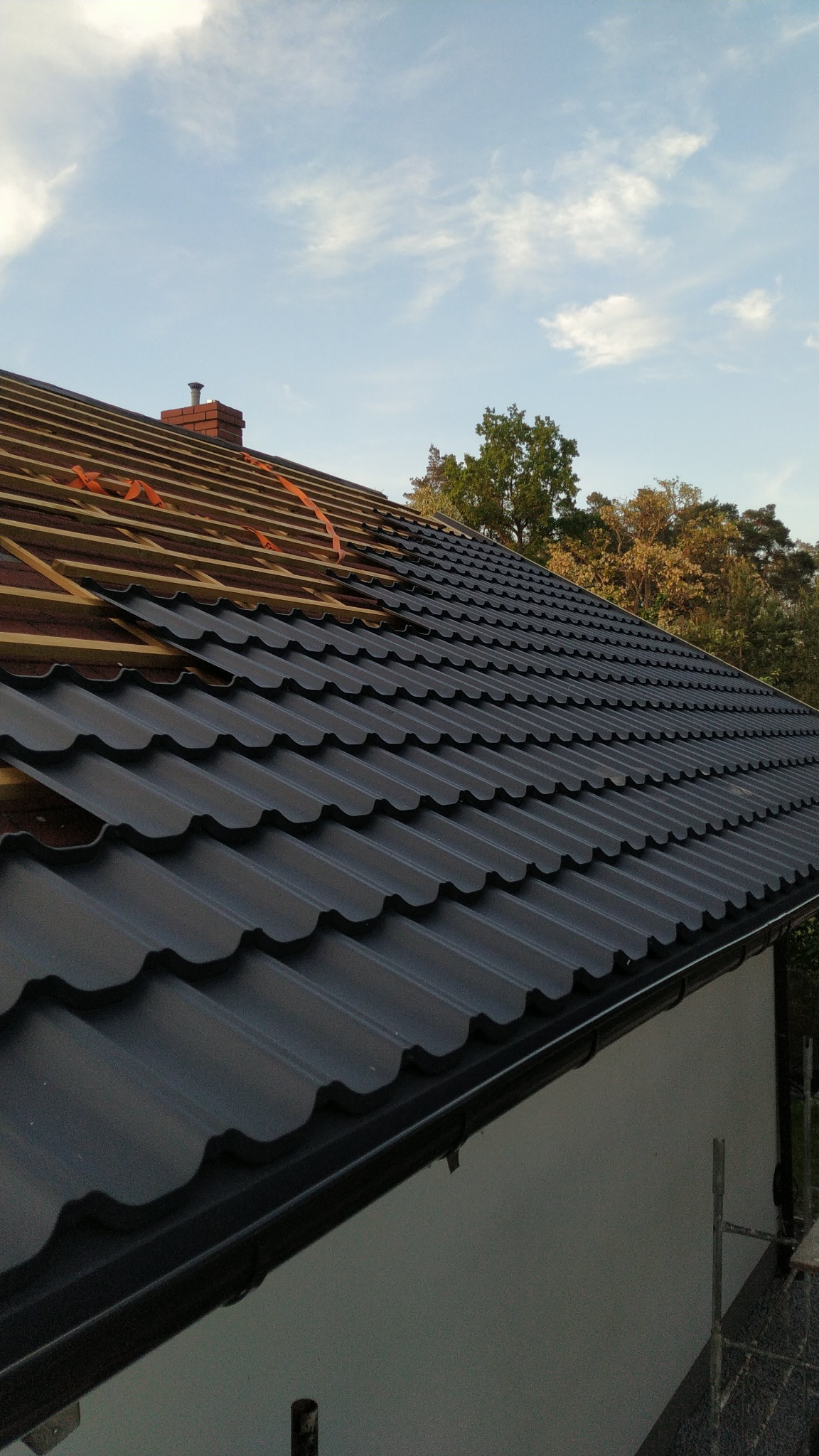 Częściowo pokryty dach z czarnej blachy trapezowej, widoczne drewniane łaty i komin na tle błękitnego nieba z chmurami i zieleni drzew.