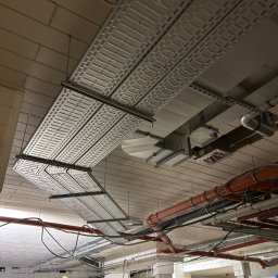 LUMARO - Montaż korytek kablowych pod sufitem w surowym wnętrzu. Widoczne rury wentylacyjne i instalacyjne. Pracownicy na drabinie i przy wejściu.