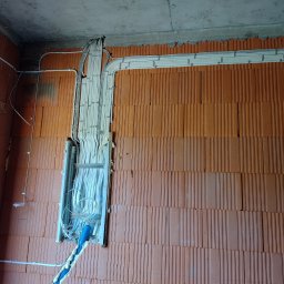 LUMARO - Instalacja elektryczna w trakcie montażu w nowym budynku z cegły. Widoczne kable w białych rurkach przymocowane do ściany i sufitu. Niebieska rura karbowana.