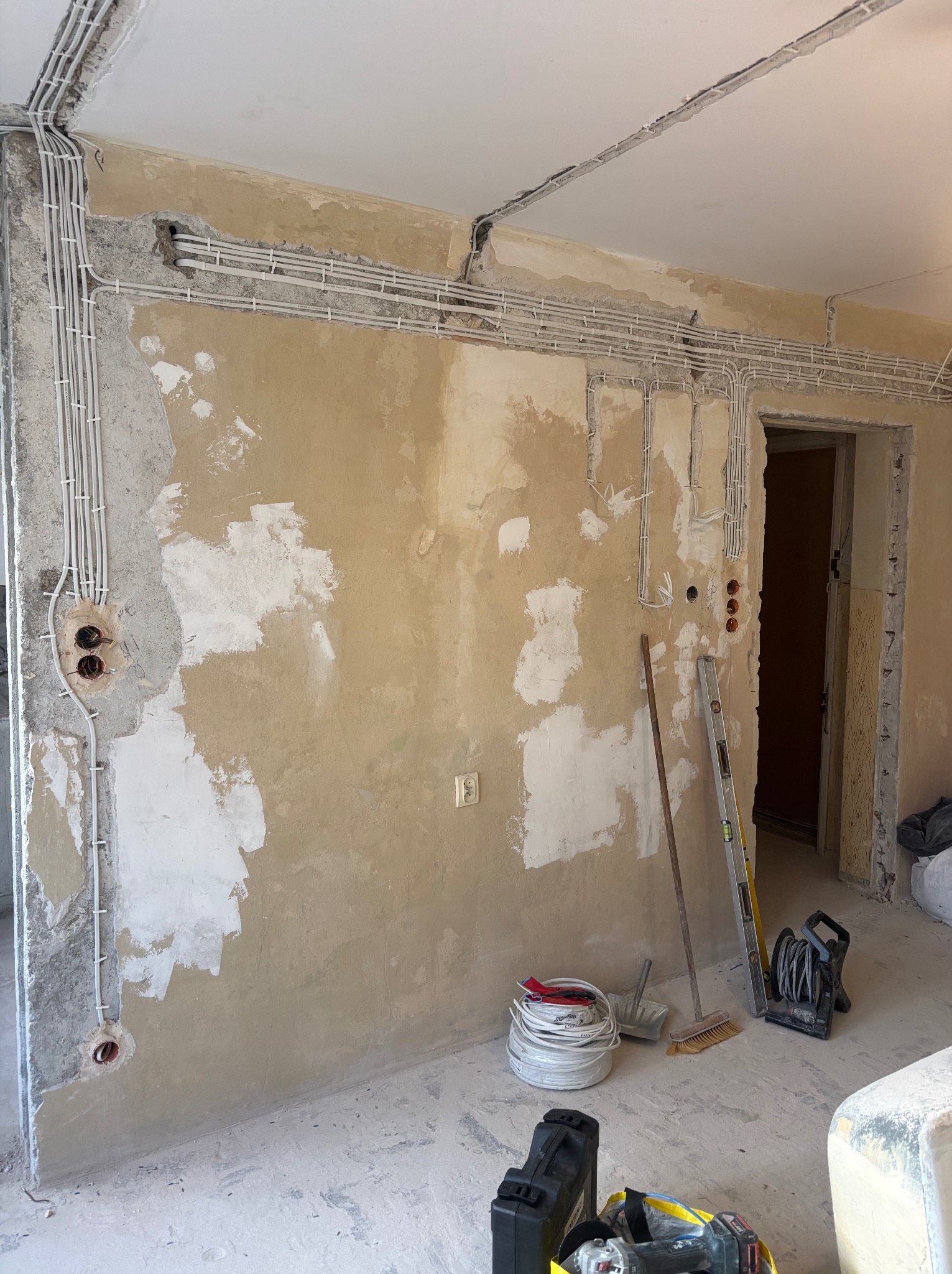 Ściana w trakcie remontu z widoczną instalacją elektryczną w białych rurkach, poziomica oparta o ścianę, narzędzia na podłodze. Widoczne puszki elektryczne.